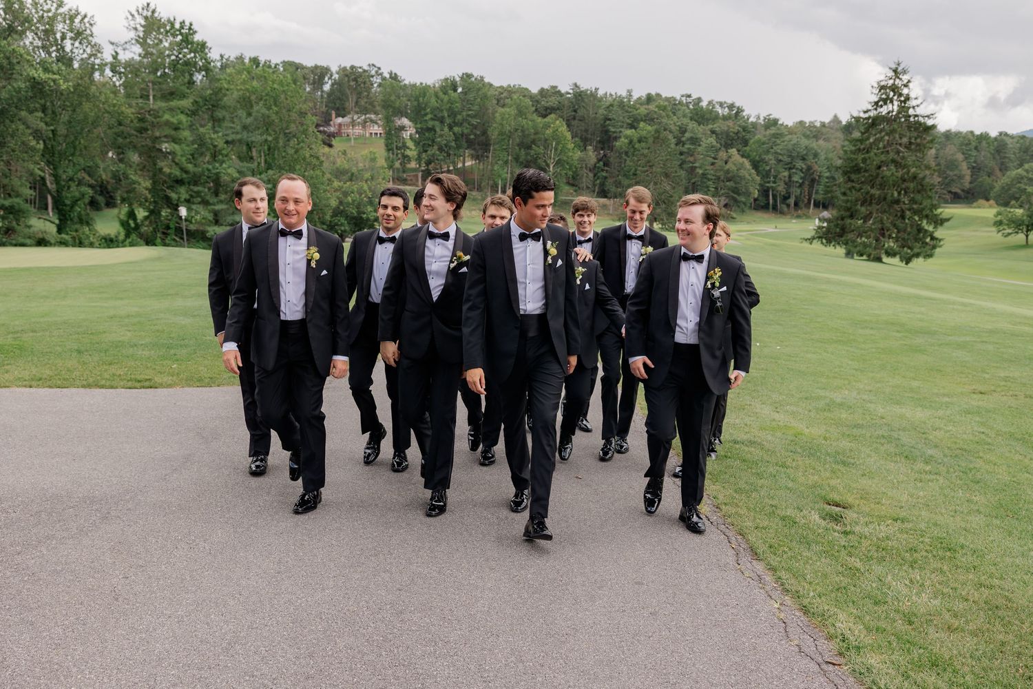 Groom and groomsmen in black tuxedos with bow ties walk confidently on a path at an outdoor wedding venue.