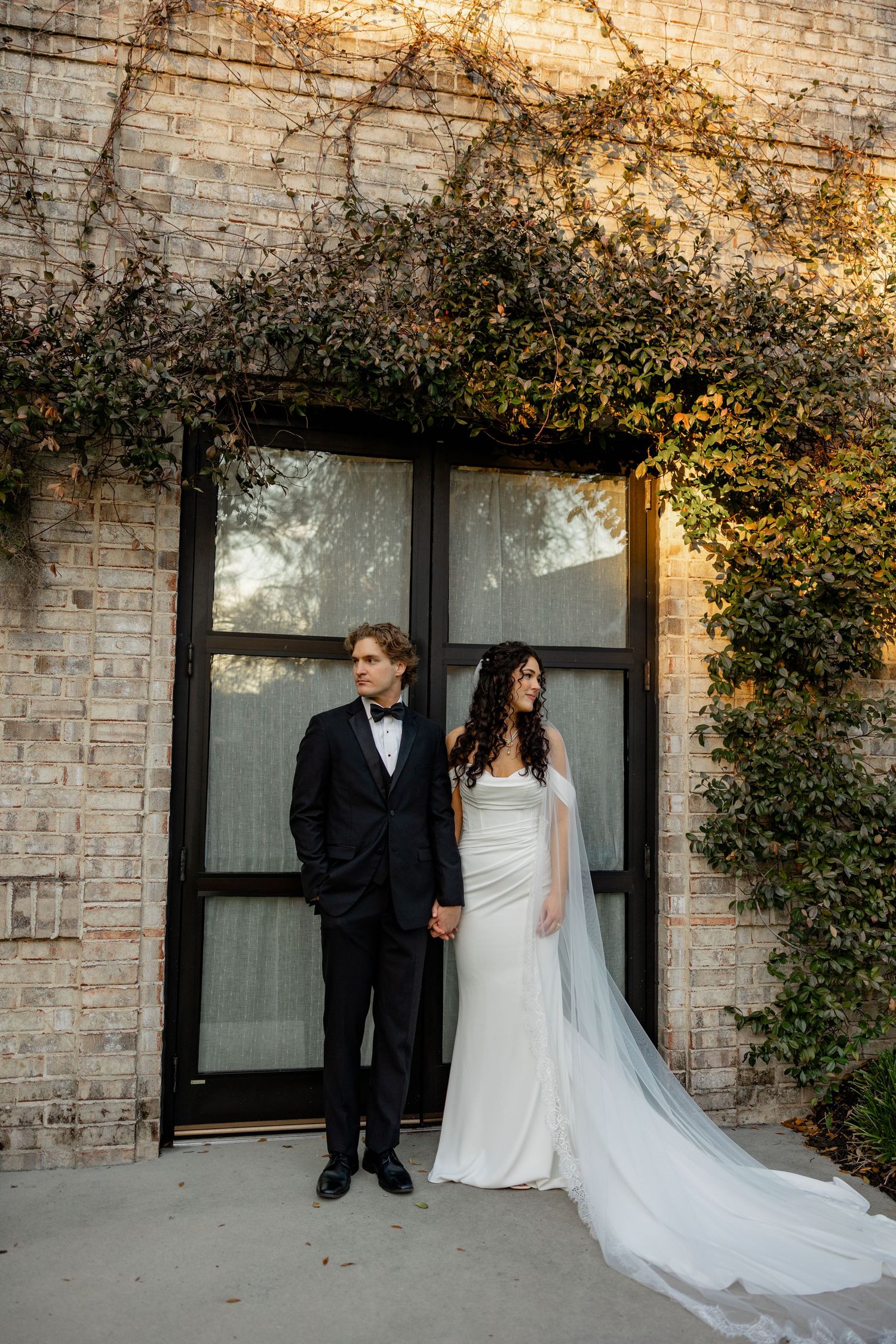 A bride and groom pose against a brick wall with climbing vines and large windows.