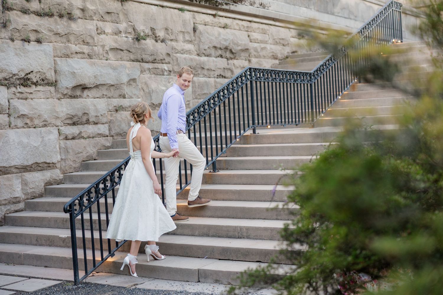 A couple in formal wedding attire descends stone steps with black metal railings in an outdoor setting.