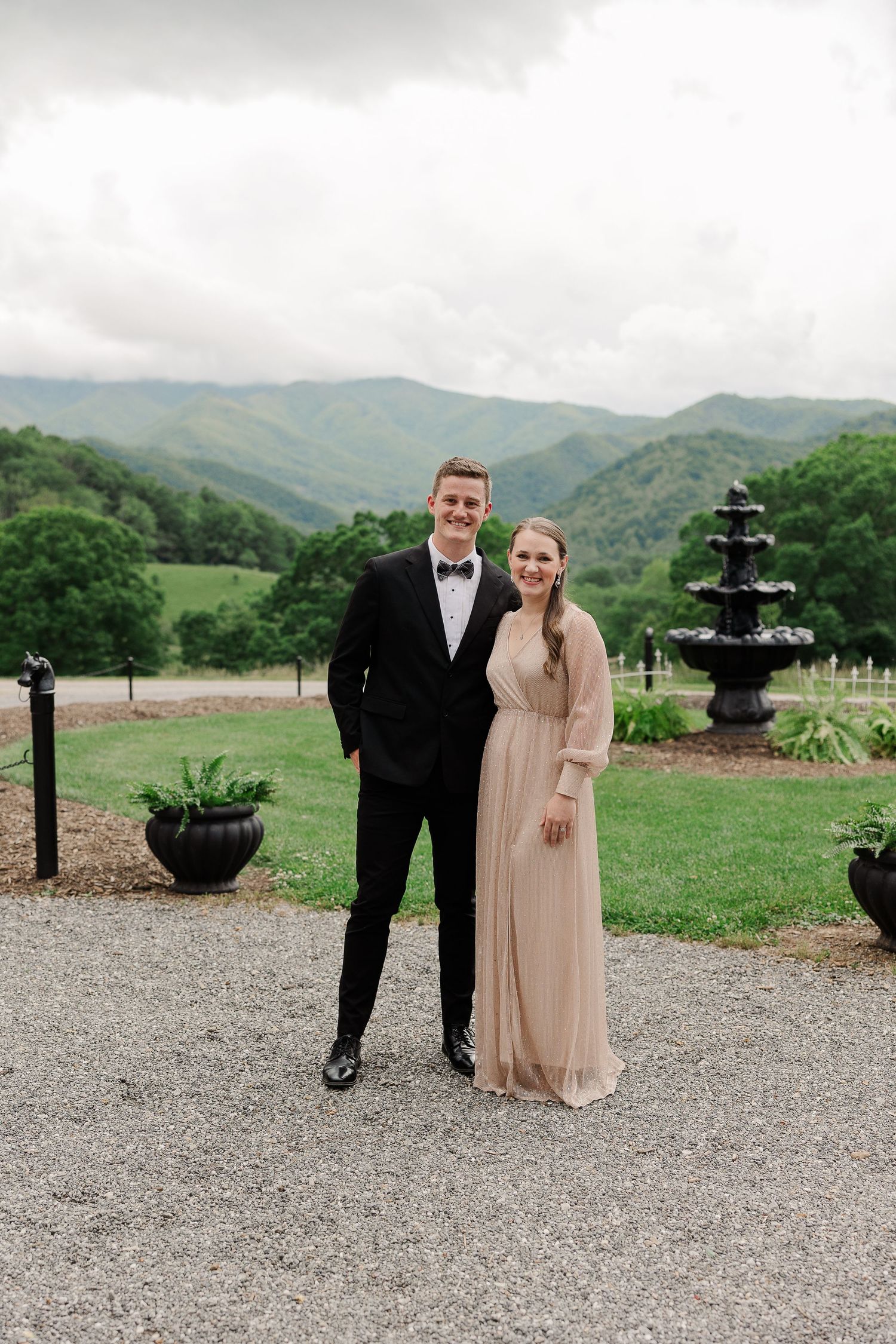 A couple in formal attire poses together on a gravel path with mountain views and a decorative fountain in the background.