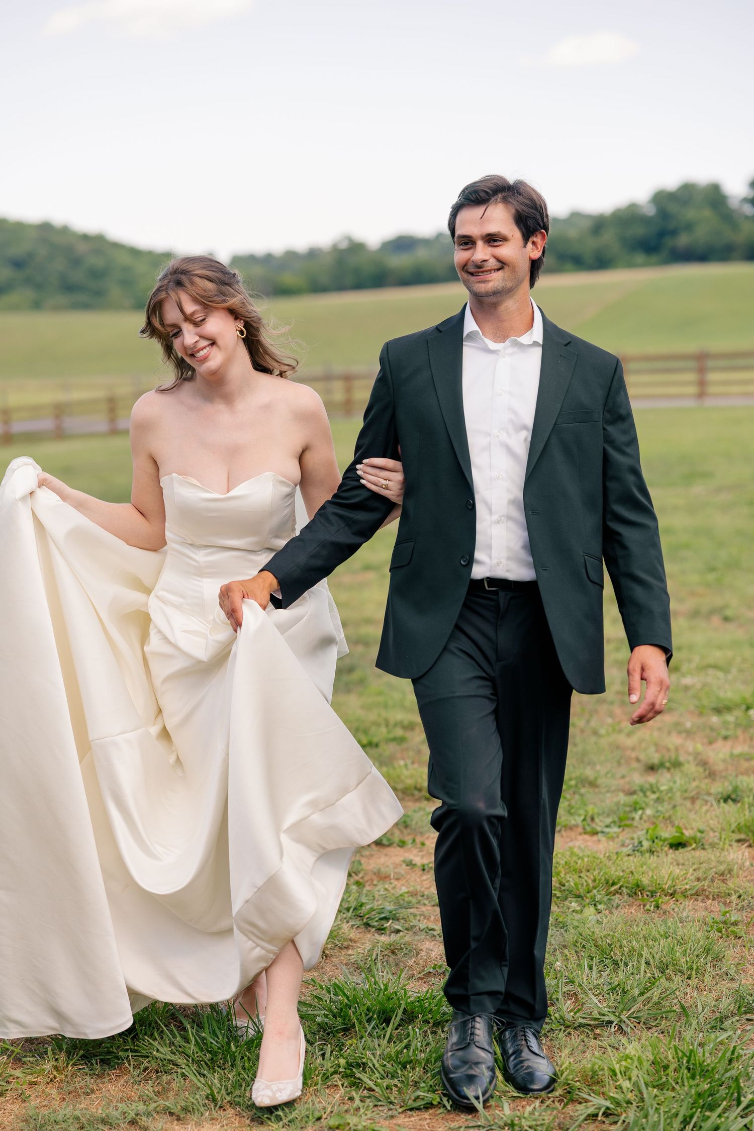 Happy bride and groom walk arm in arm through a scenic green countryside field after their wedding.