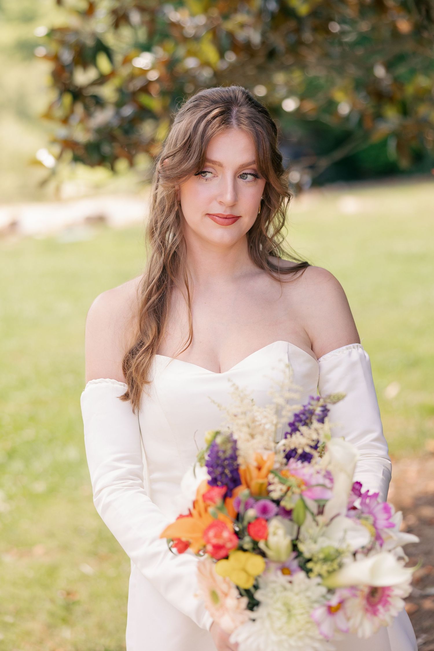Elegant bride in off-shoulder gown holds vibrant wildflower bouquet in a sunlit garden.