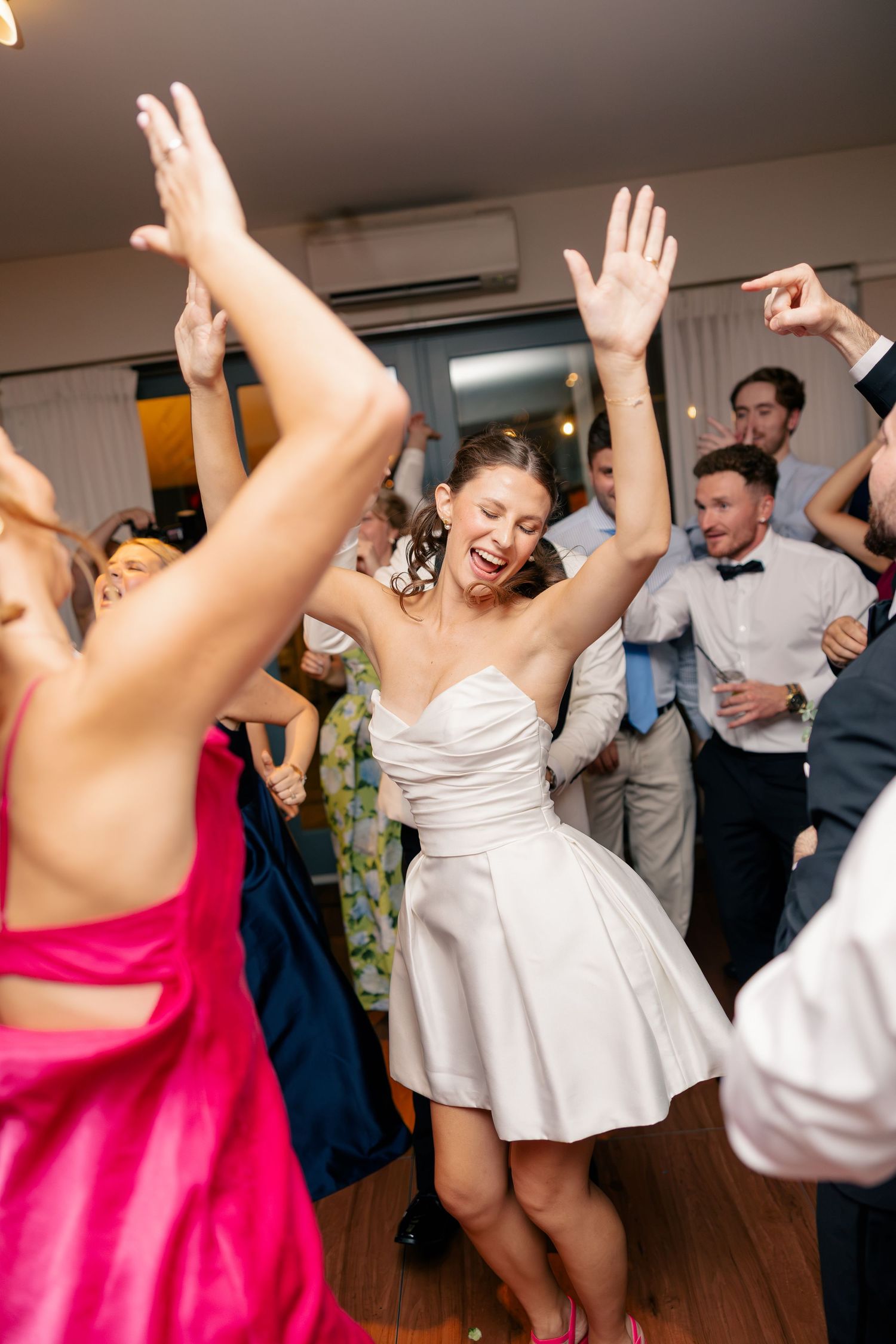 Bride in short white dress dances joyfully with raised arms surrounded by guests at wedding reception.