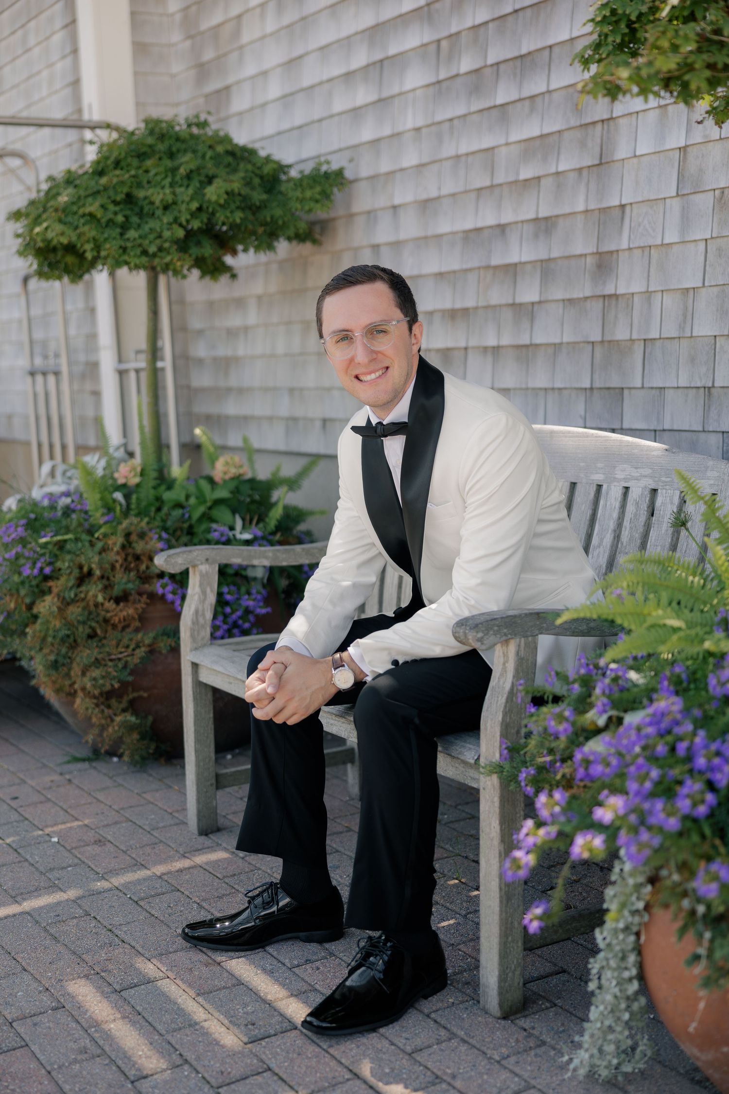 Dapper groom in white tuxedo jacket and black bow tie sits on outdoor bench surrounded by flowers.