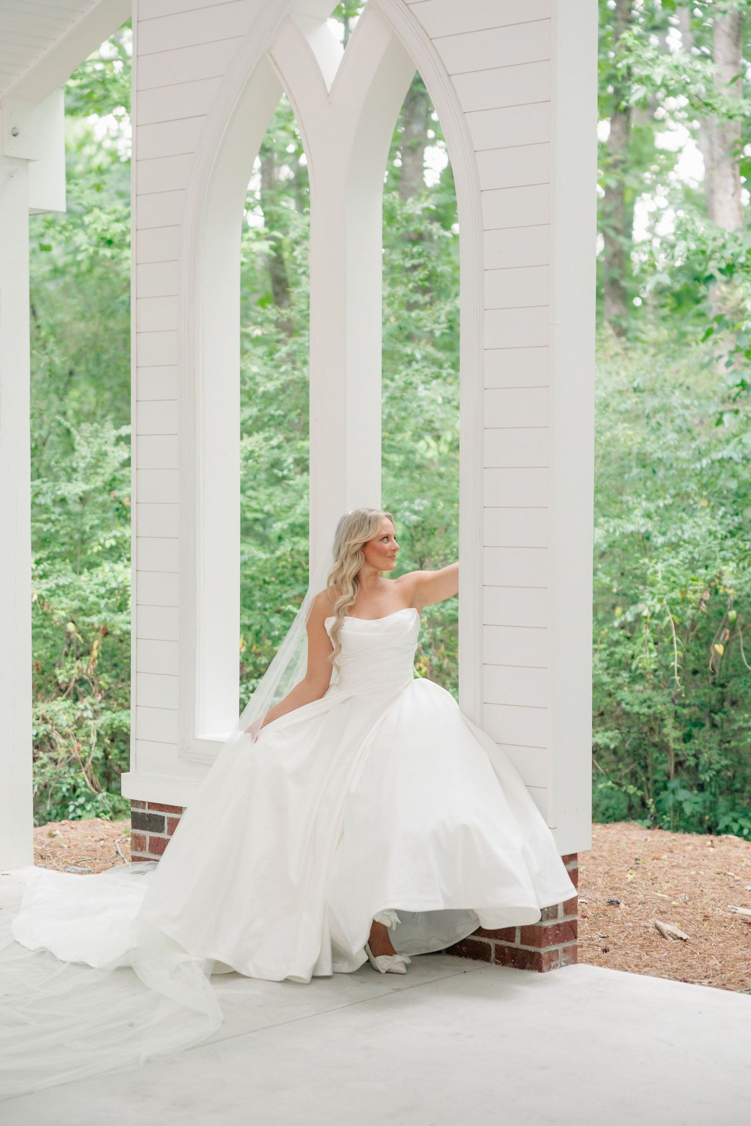 Bride in strapless white ballgown with veil poses under white arched chapel columns outdoors.