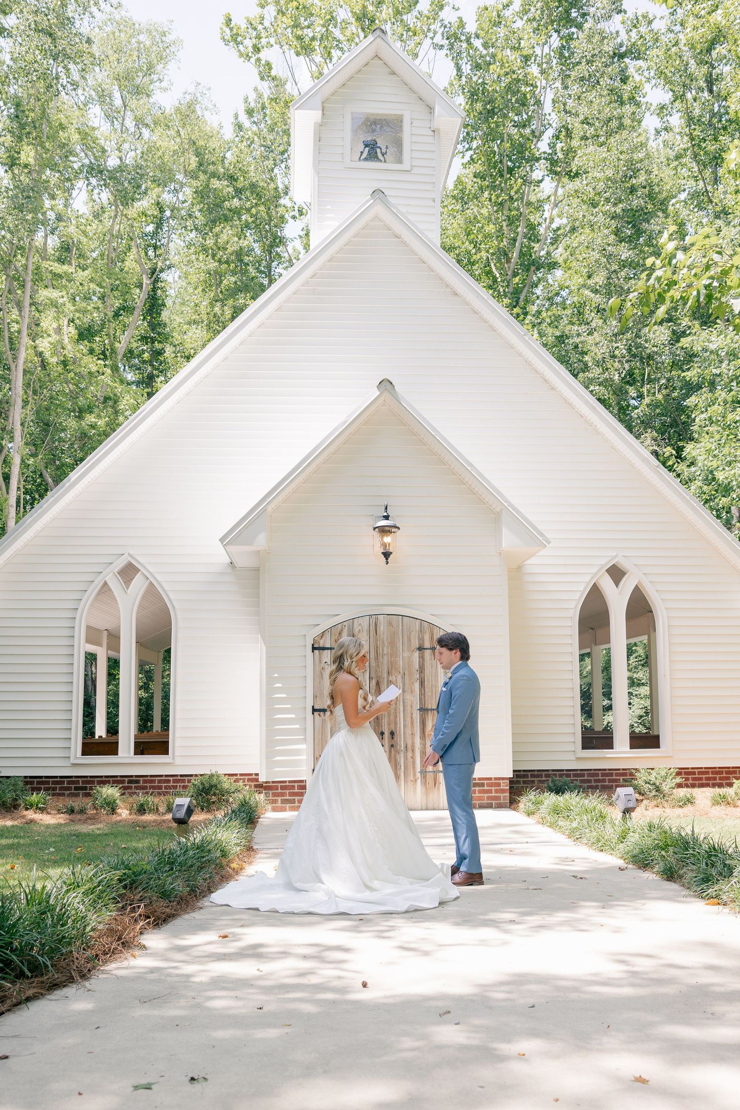 A bride and groom exchange vows outside a charming white chapel surrounded by lush green trees.