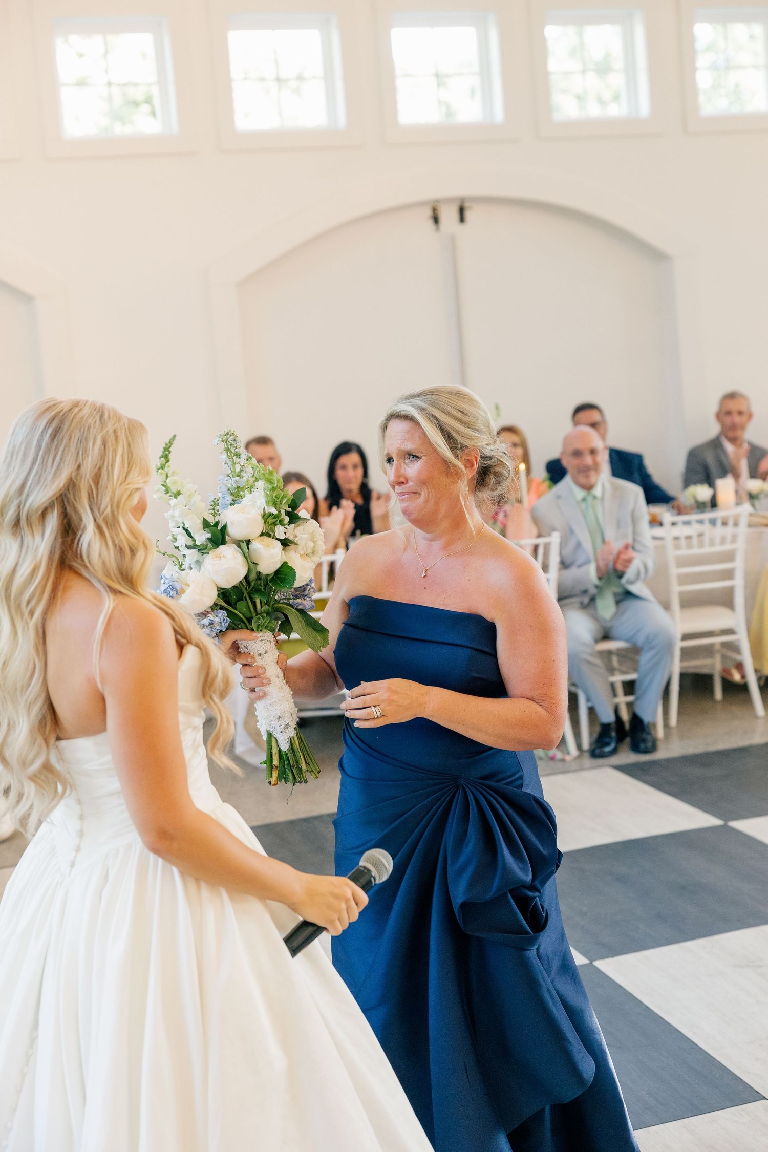 Bride in white gown receives her bouquet from a woman in a navy strapless dress during reception.