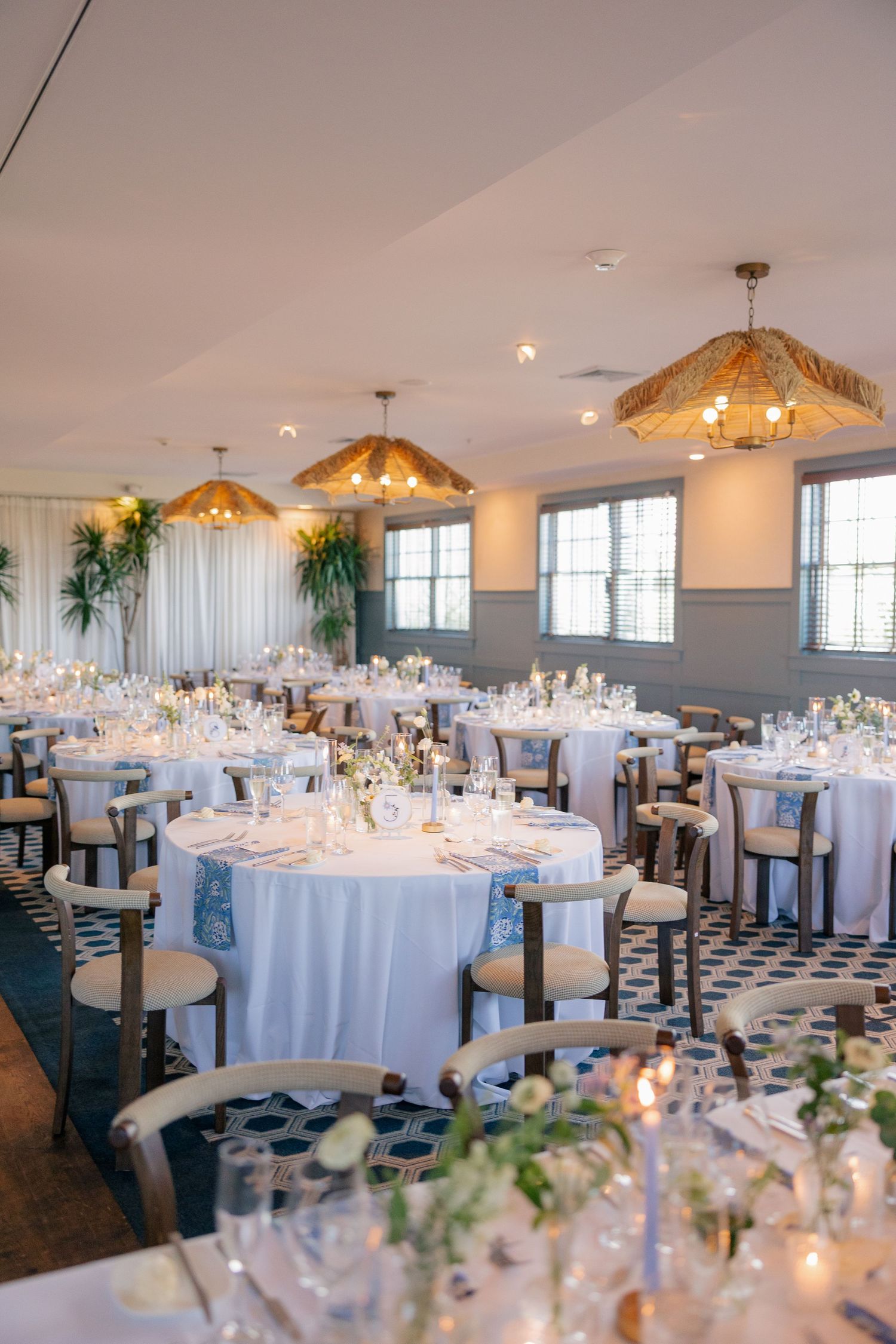 Elegant wedding reception hall with round tables, white linens, rattan pendant lights, and candles.