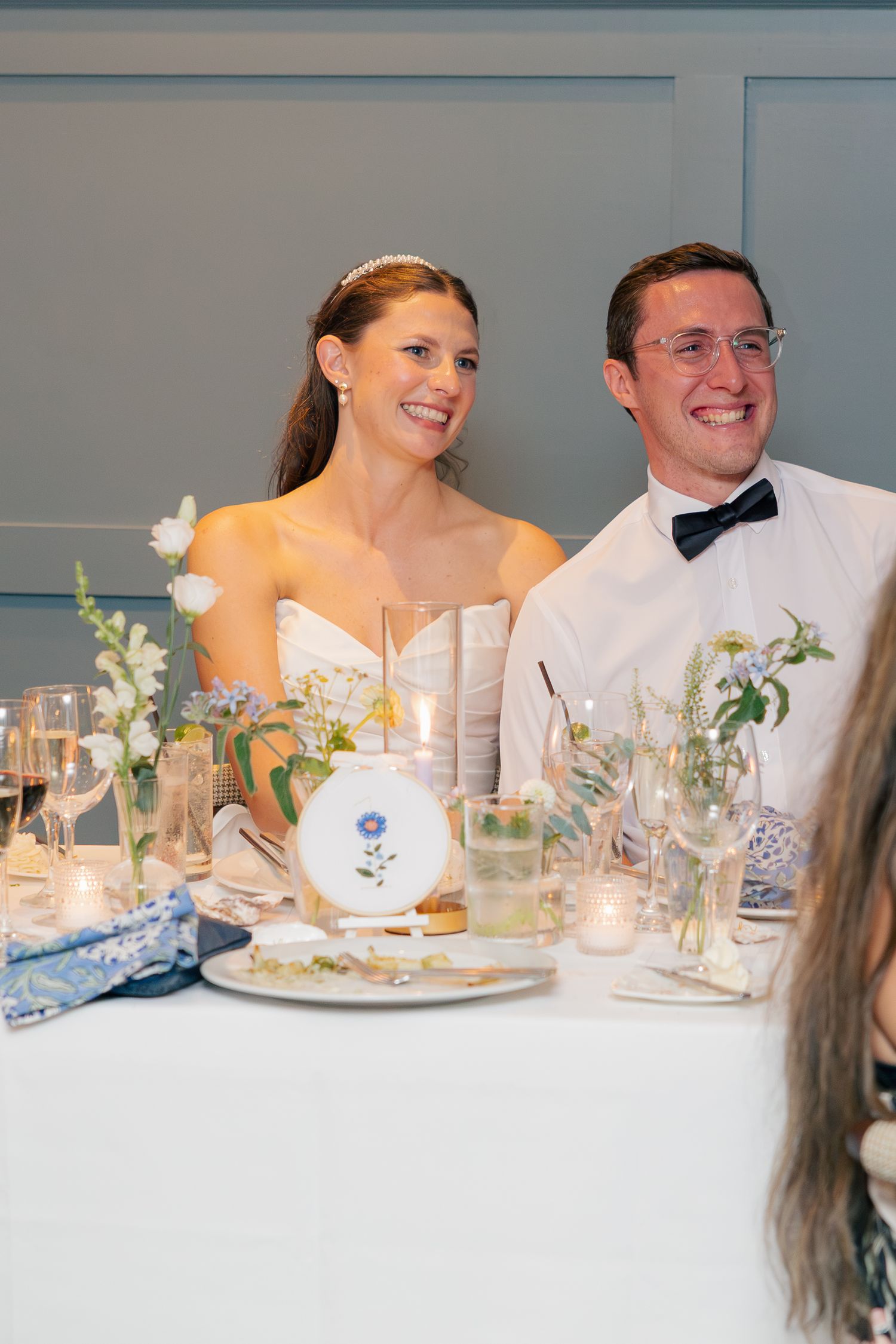 Bride and groom smiling at wedding reception table with floral centerpieces, candles, and elegant blue table settings.