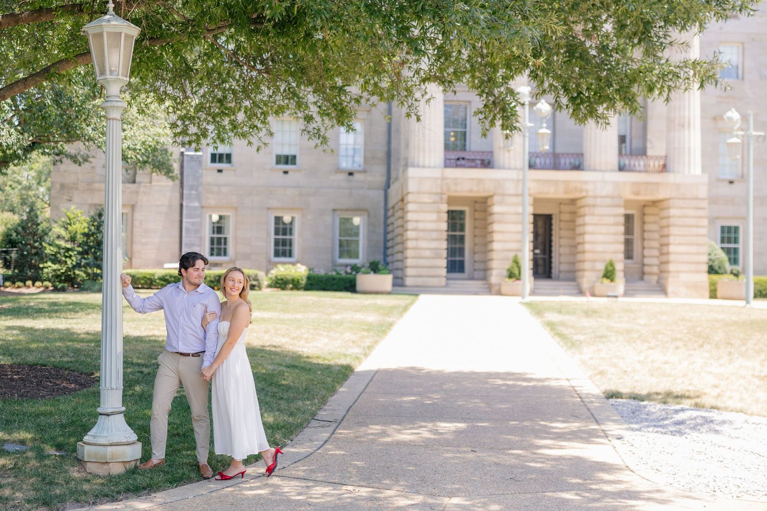 Engaged couple in white dress and khakis pose by lamp post outside grand stone building on sunny day.