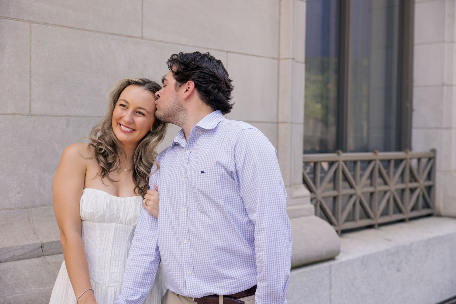 A man in a blue shirt kisses a smiling woman in a white strapless dress on the cheek outdoors.