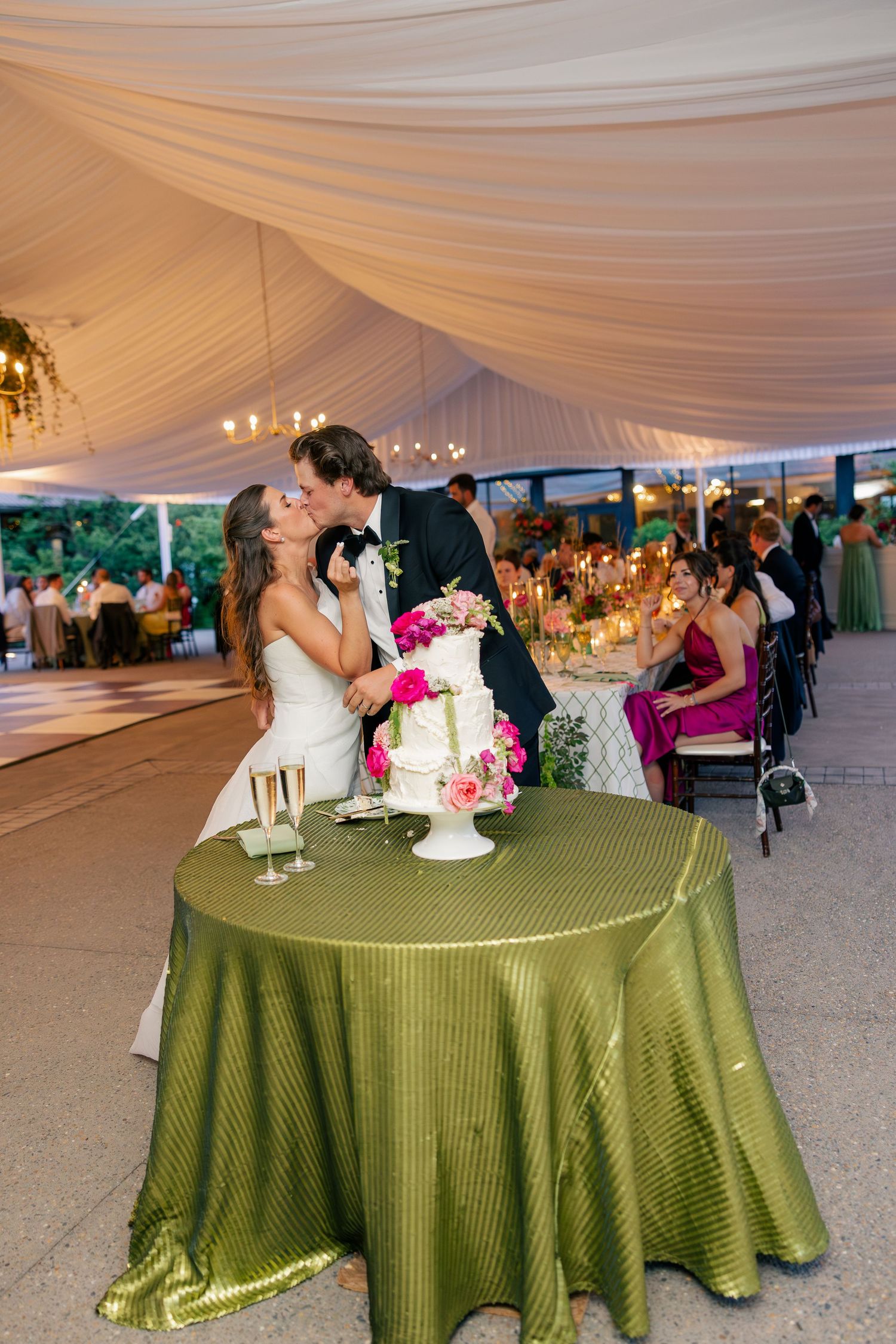 Bride and groom share a kiss at wedding cake table with green tablecloth under elegant draped tent reception.