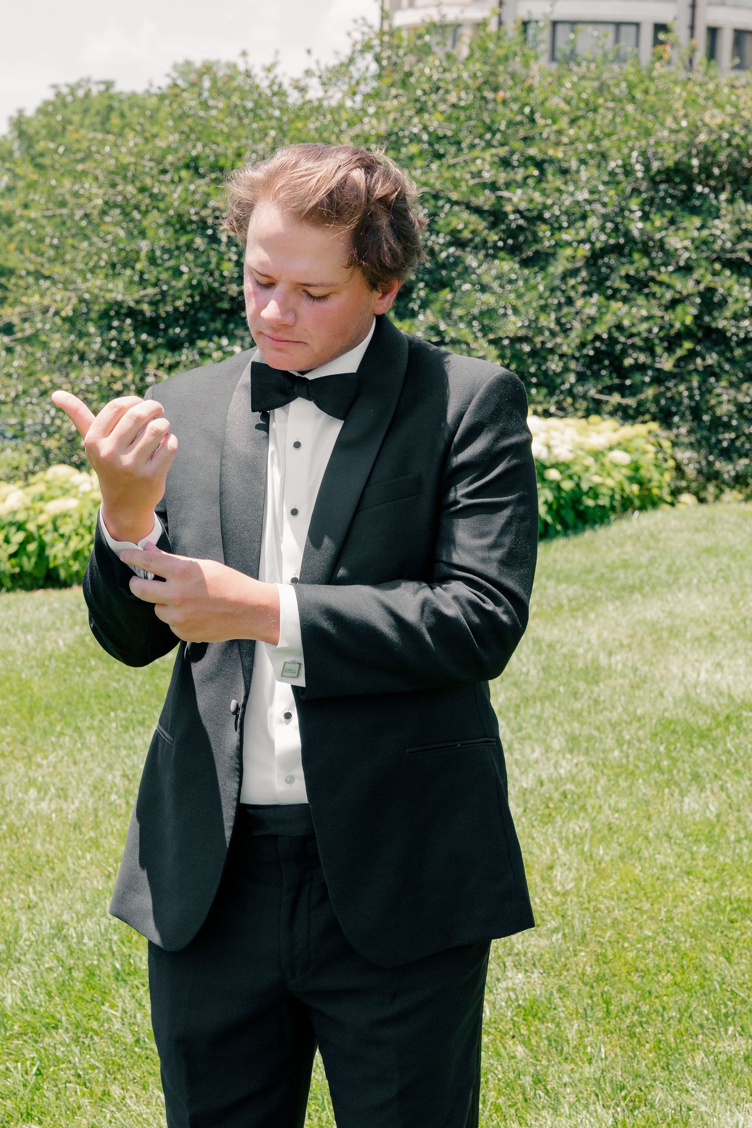 Groom in black tuxedo and bow tie adjusting his cufflinks outdoors on a sunny wedding day.