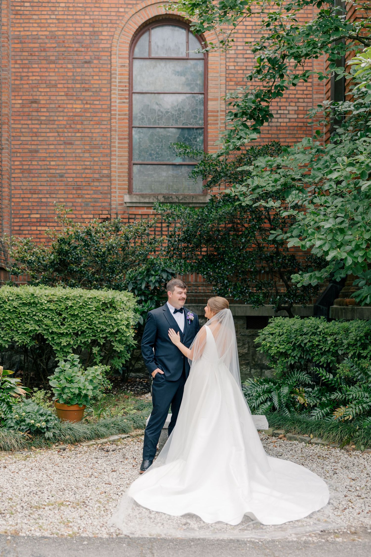 Bride in flowing white gown and groom in navy suit pose in church garden.