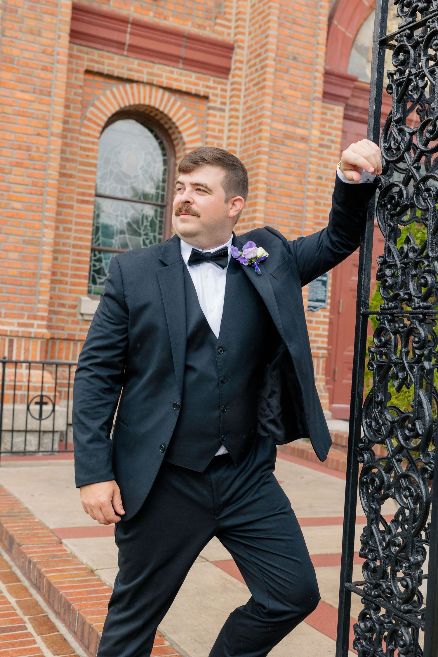 Dapper groom in black tuxedo with bow tie and boutonniere posing by ornate iron gate at brick church.