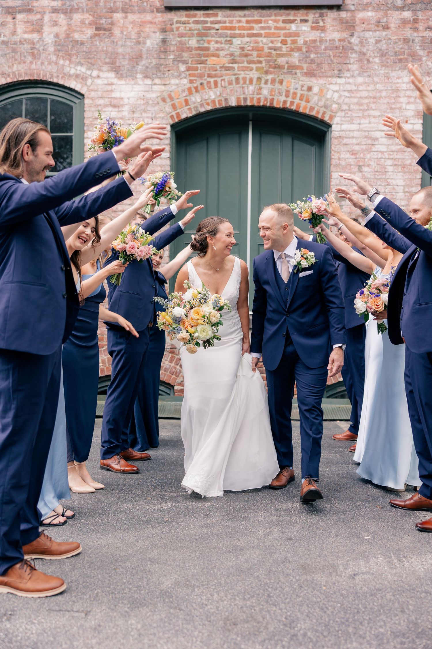 Wedding party forms a celebratory arch for the bride and groom outside a rustic brick venue.