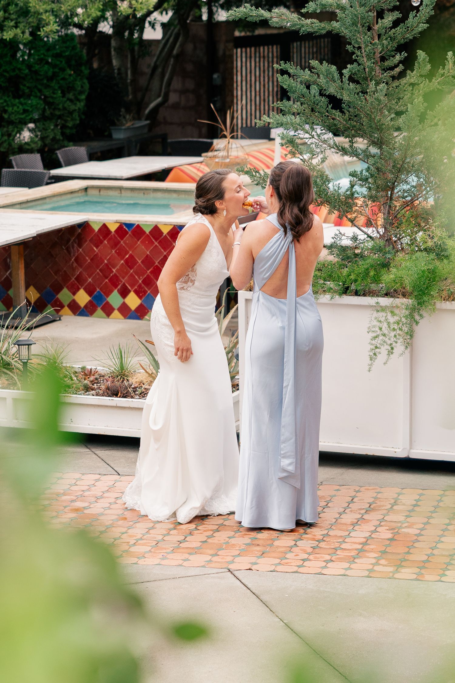Bride in white gown and bridesmaid in light blue dress sharing a moment near a pool patio garden.