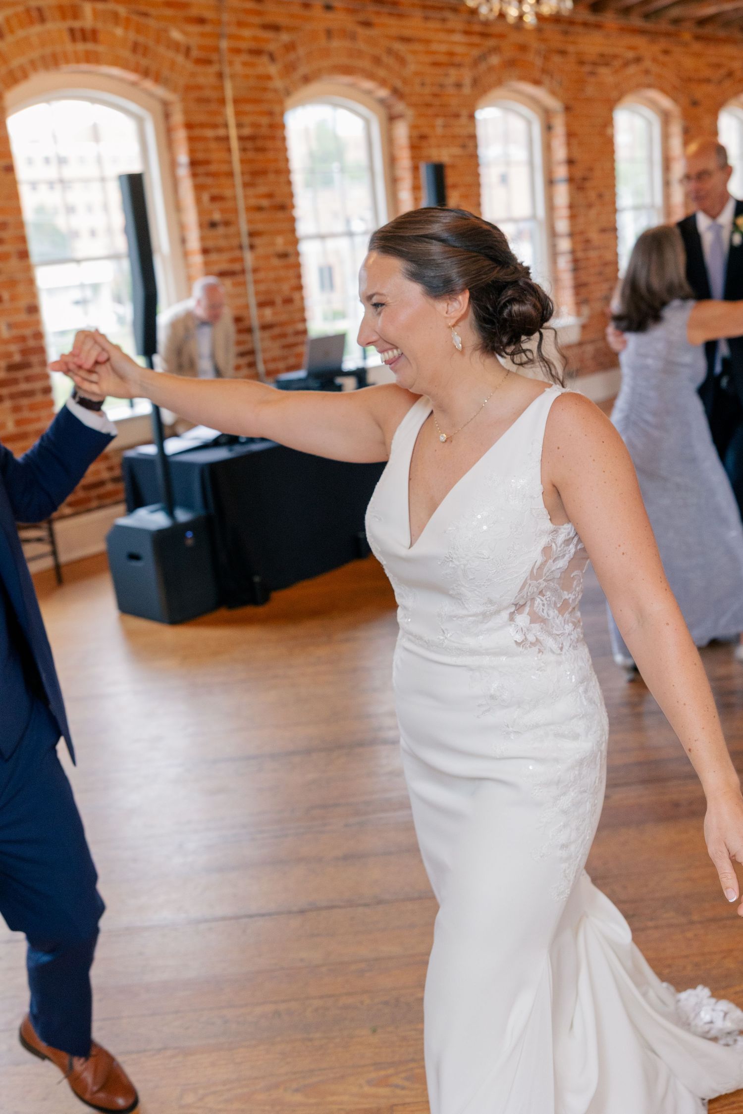 Radiant bride in lace wedding gown laughs while dancing with groom in brick-walled reception venue.