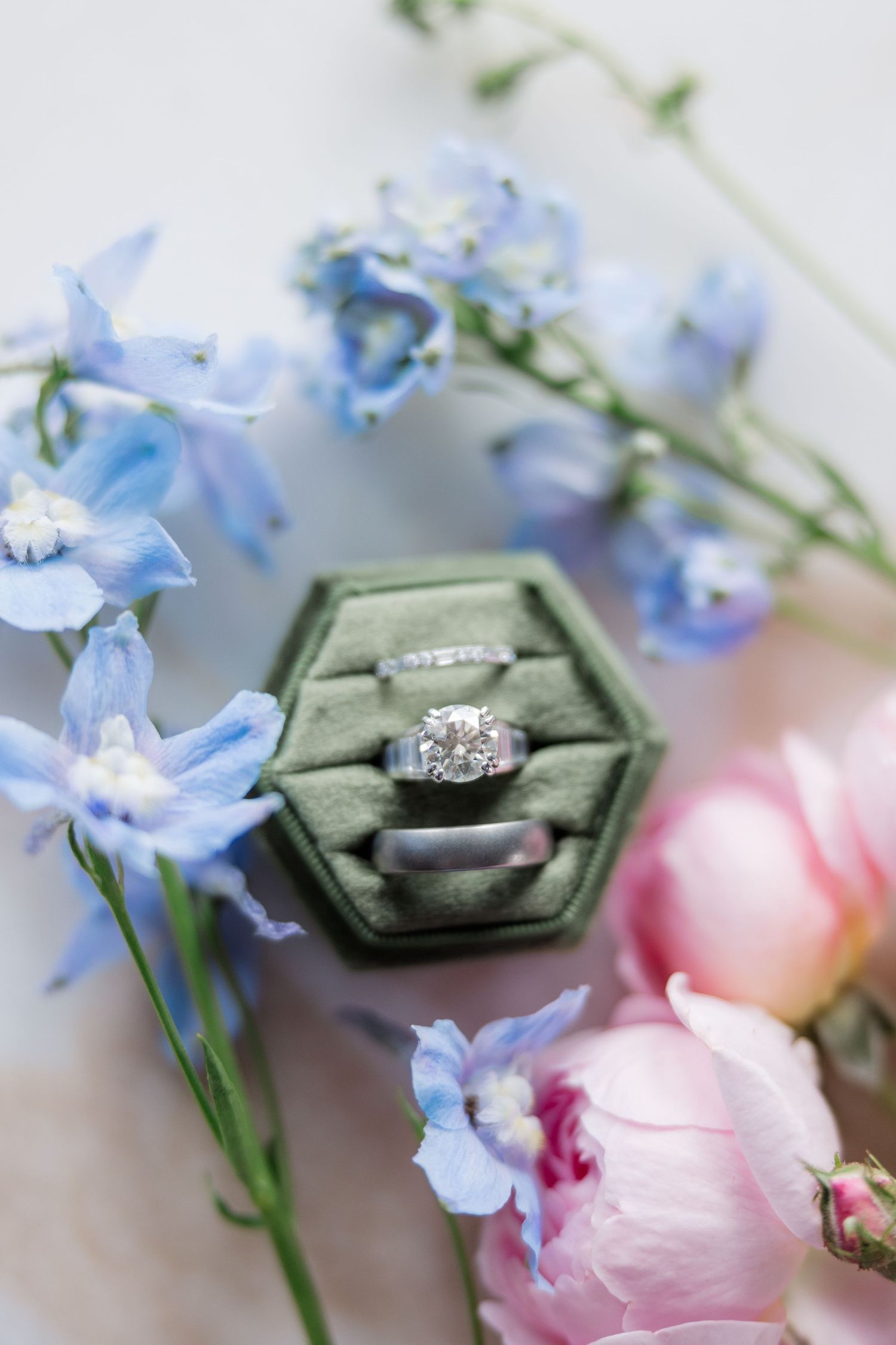 Engagement ring rests on green velvet ring box surrounded by delicate blue and pink flowers.