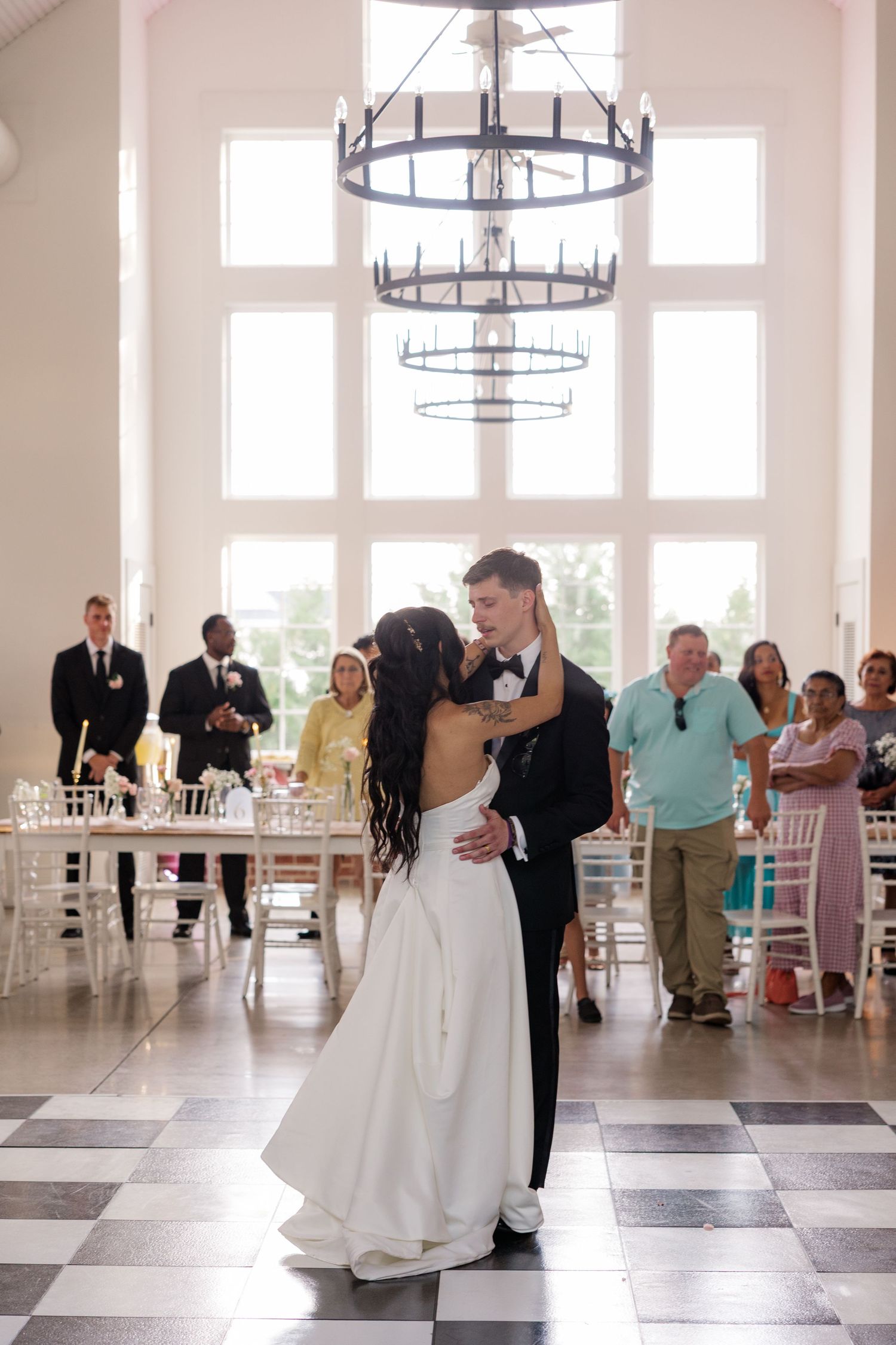 Wedding couple dances in a bright white reception hall with chandeliers and checkered floor.