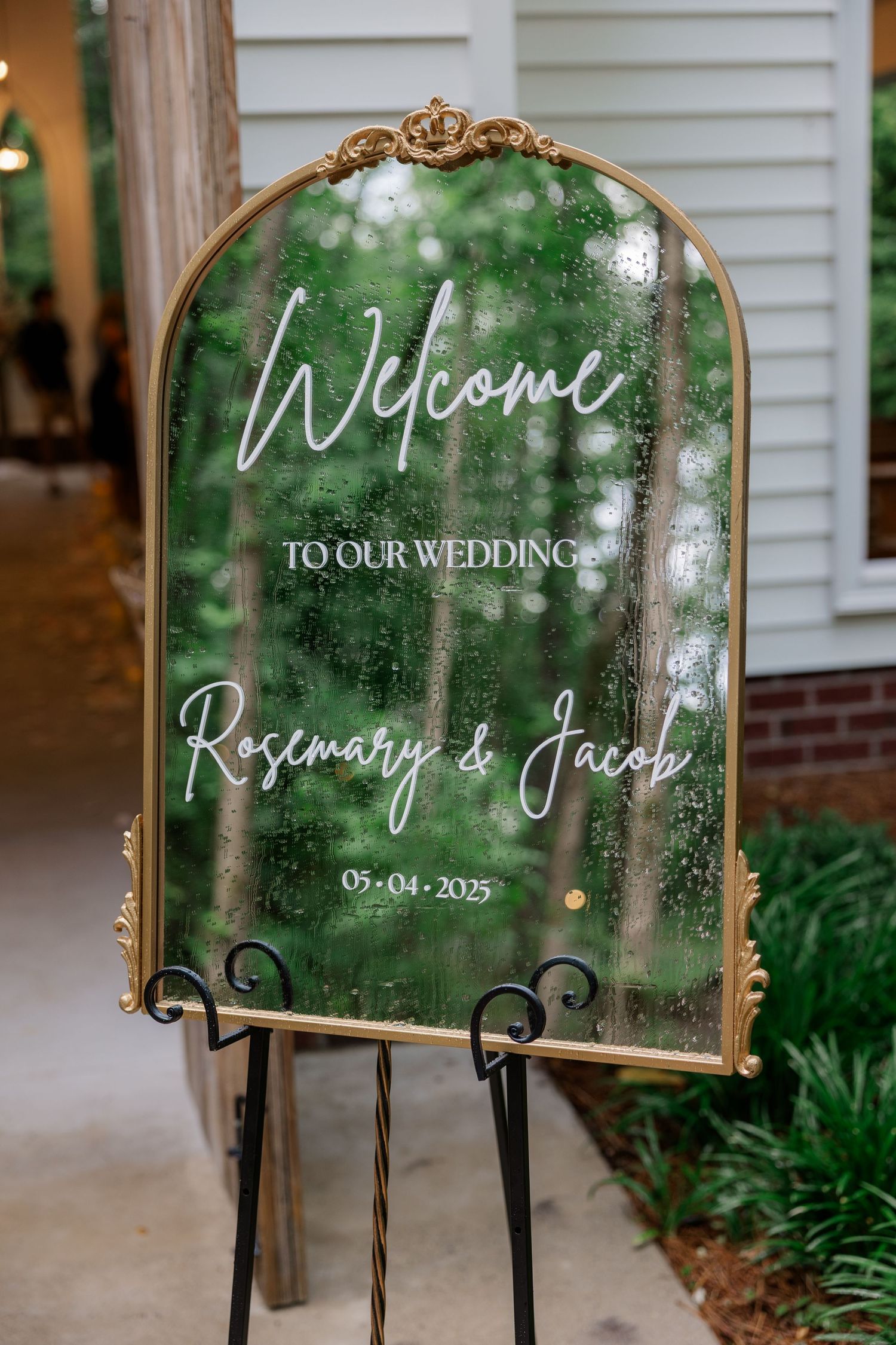 Elegant mirror welcome sign with white calligraphy displayed at outdoor wedding venue.