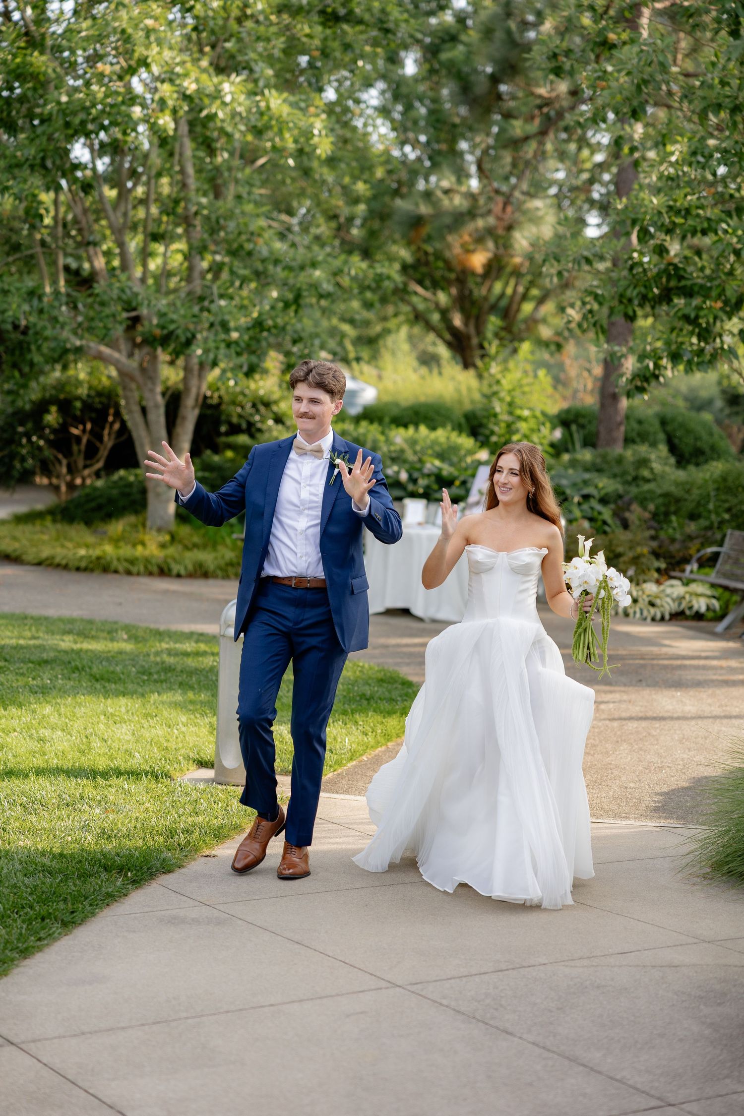 Playful wedding couple walking and posing on a garden path surrounded by lush greenery.