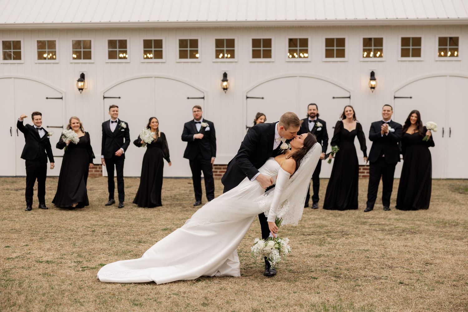 Wedding party in black attire poses creatively outdoors against white building.