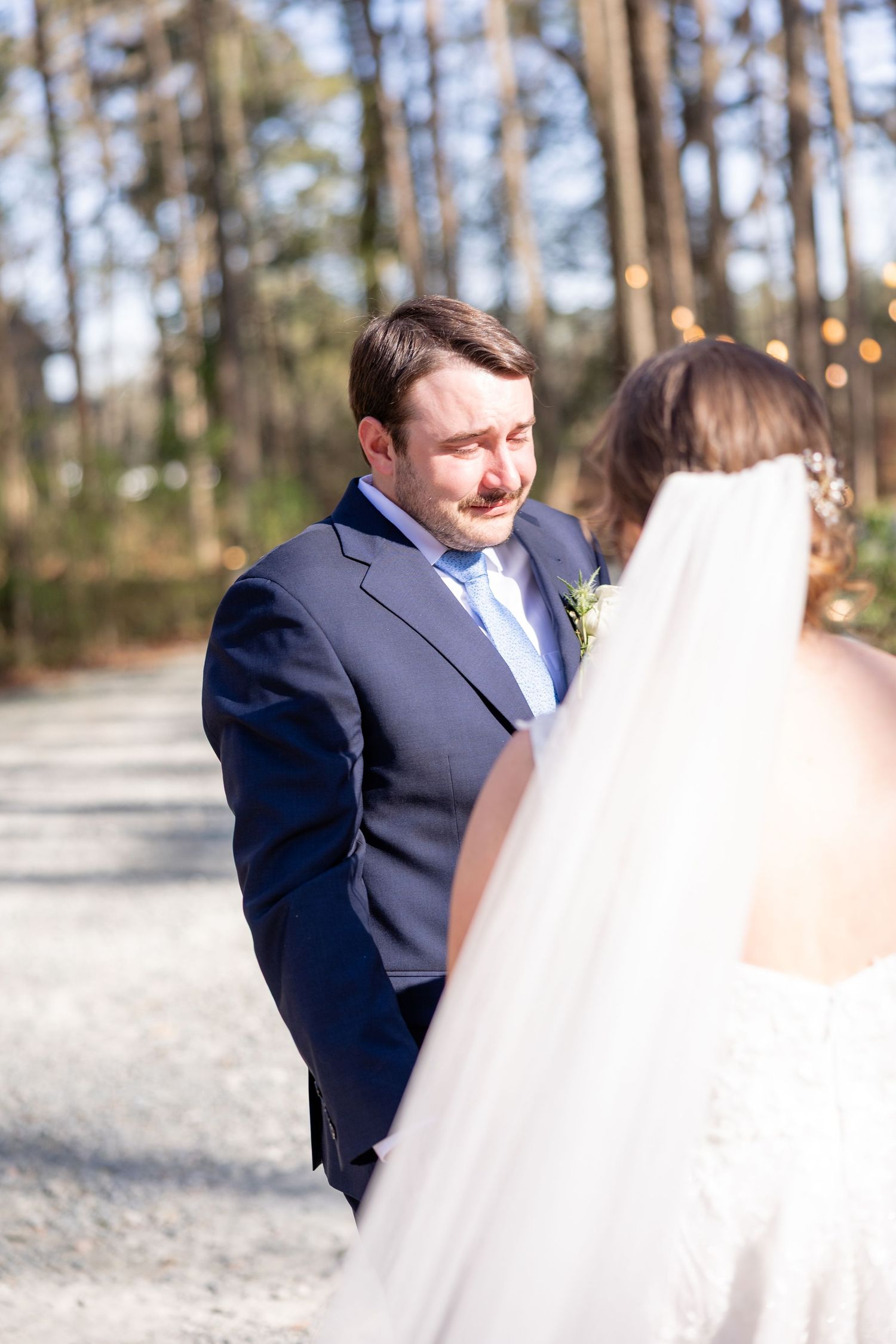 First look wedding moment captured between couple in navy suit and white gown among tall pine trees.