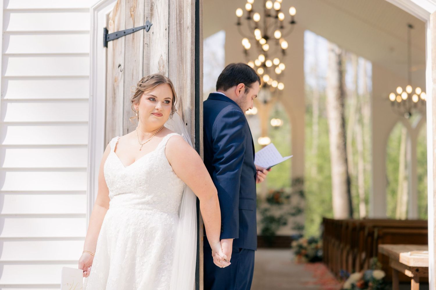 Wedding party members gather outside white-sided venue with chandelier visible.
