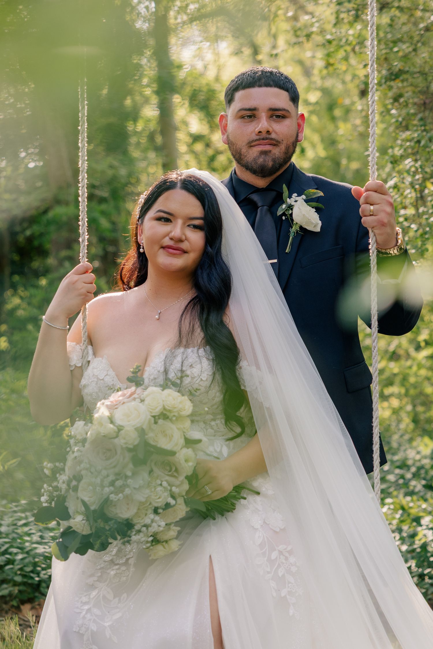 A romantic couple posing on a rustic swing in a sun-dappled forest setting on their wedding day.