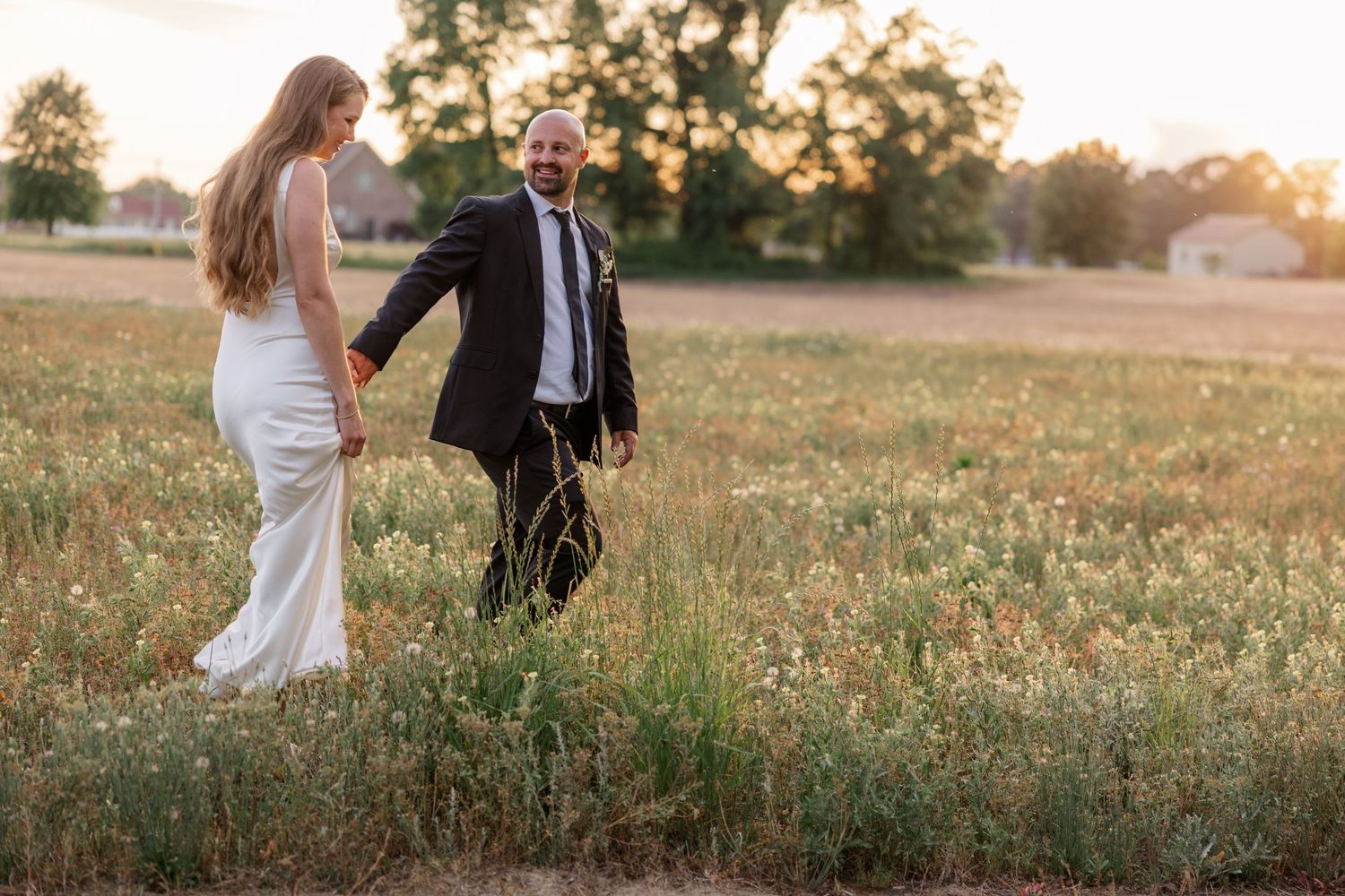 Newlyweds walking through a golden field at sunset holding hands in formal wedding attire.