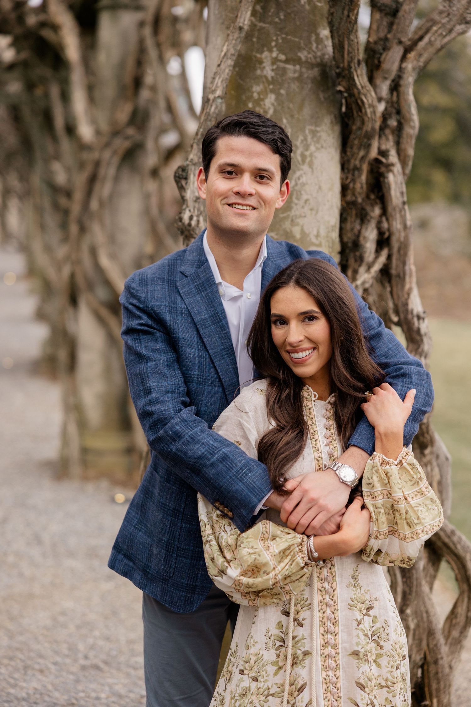 A couple embraces outdoors among trees, with the woman wearing a floral dress and the man in a navy blazer.