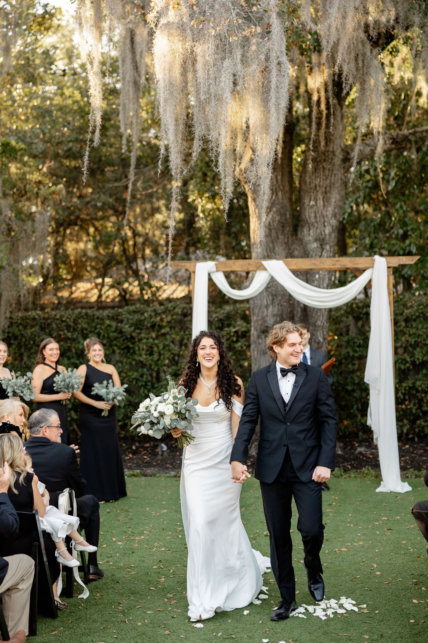 Newlyweds walking down aisle at outdoor garden wedding with Spanish moss hanging from trees above.