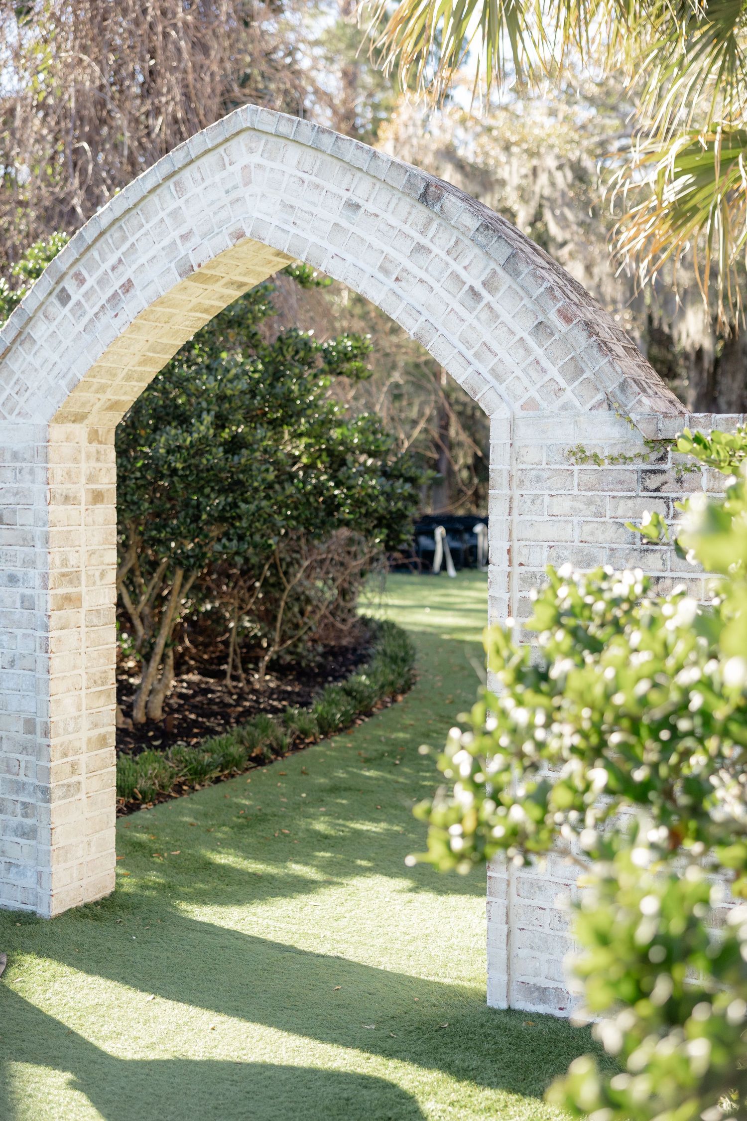 White stone archway leading through a lush garden path with palm trees and greenery.