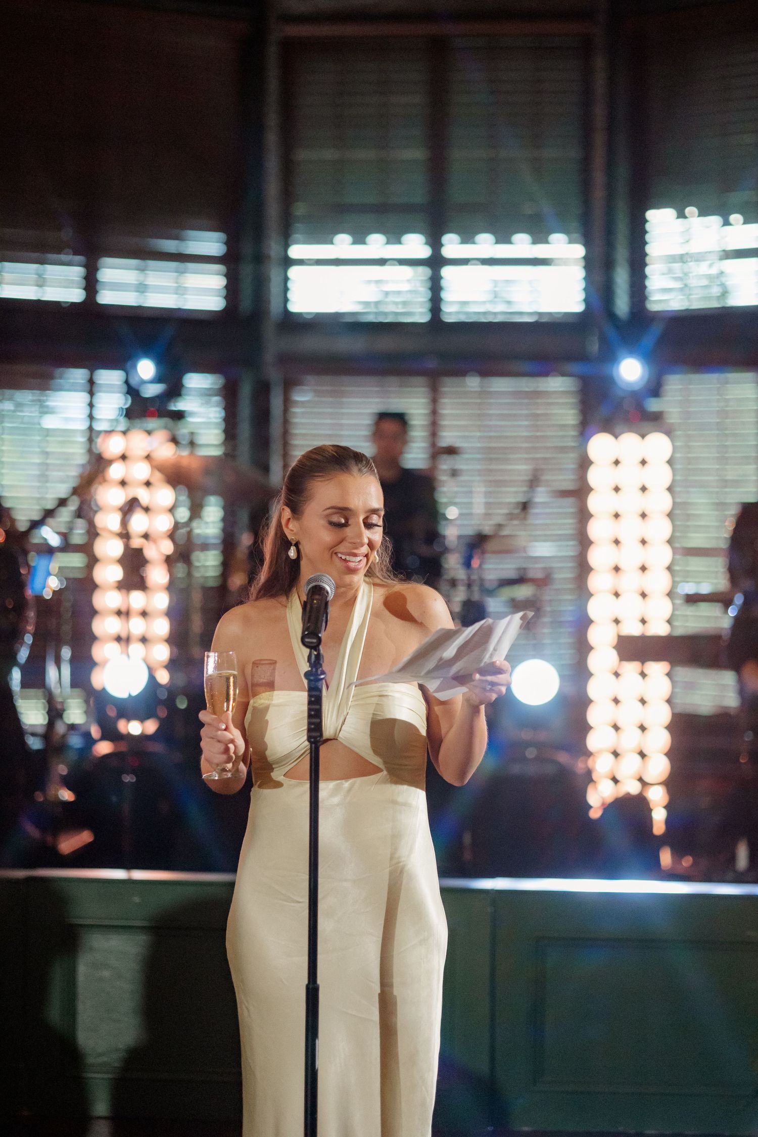 Woman in ivory halter jumpsuit gives a toast at a microphone during a wedding reception with stage lights.