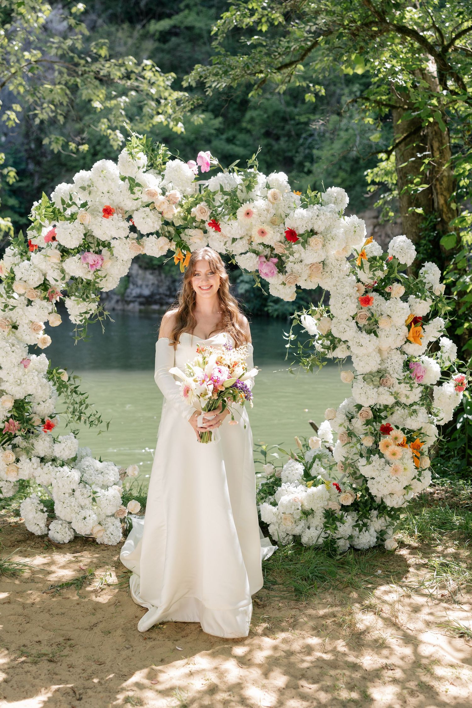 Radiant bride holding a colorful bouquet standing beneath a white floral circular arch by a river.