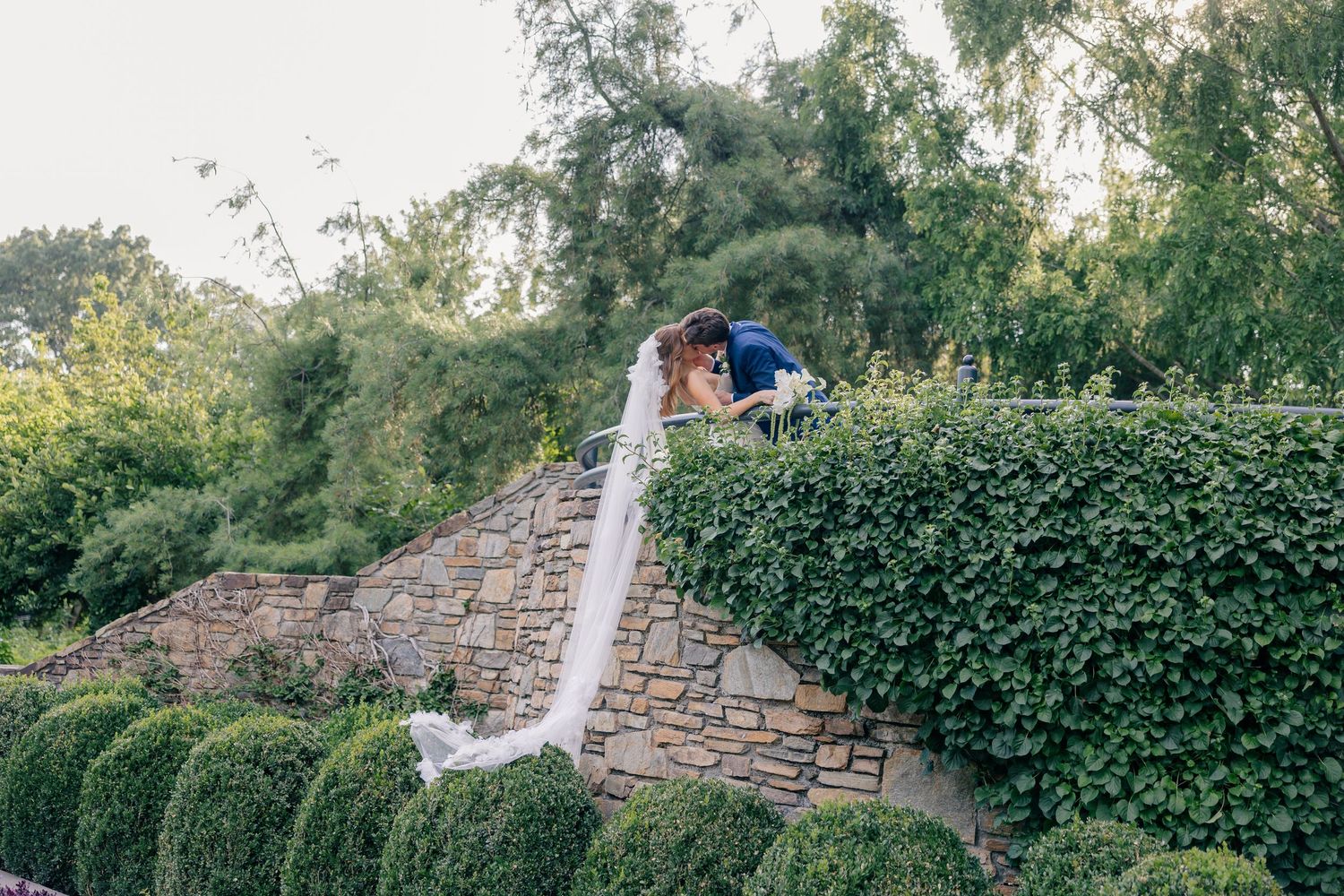 Bride and groom sharing a romantic kiss on a stone garden wall surrounded by lush green hedges and trees.
