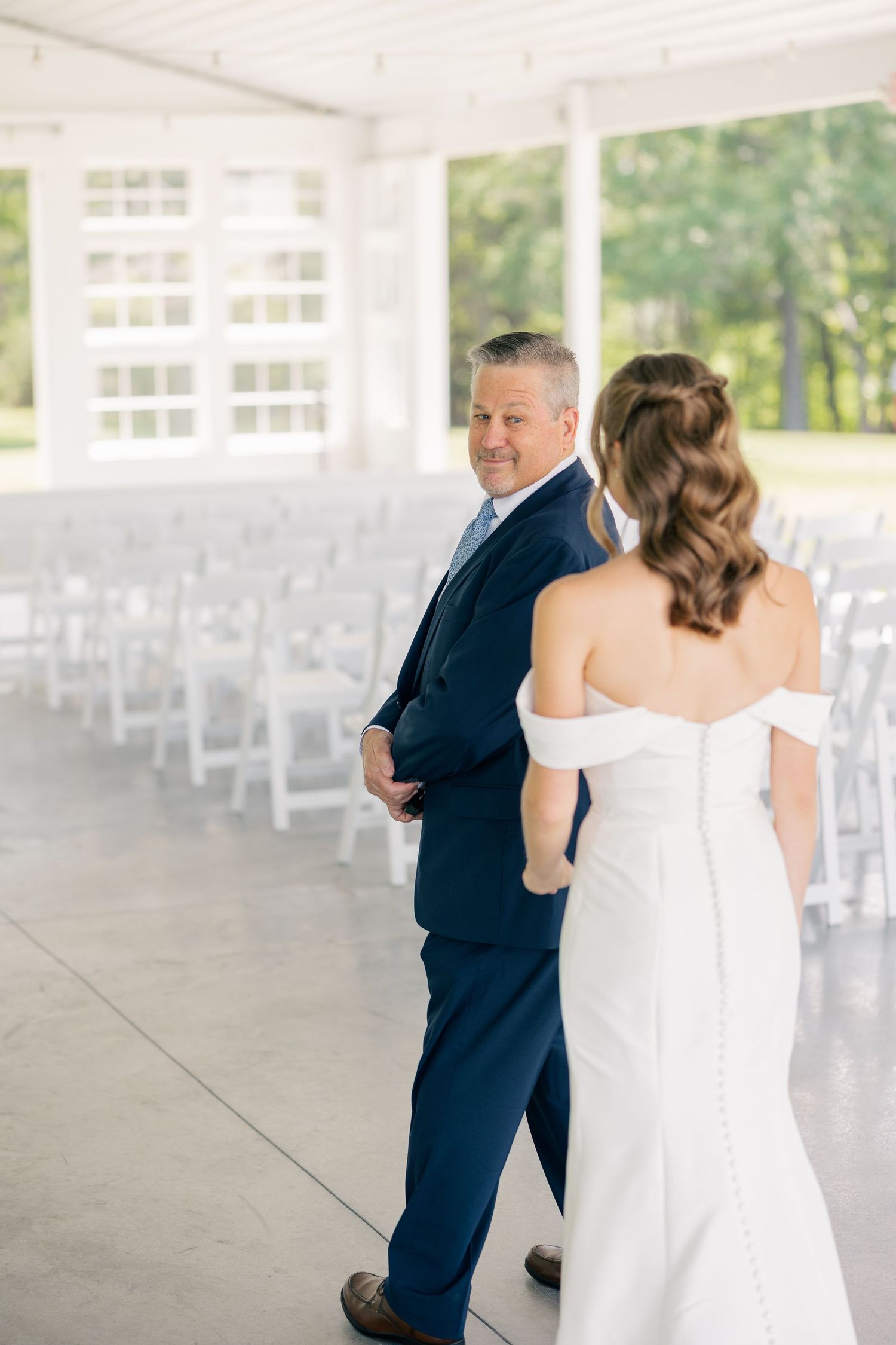 Groom in navy suit smiles at bride in off-shoulder white gown during first look in elegant white pavilion.