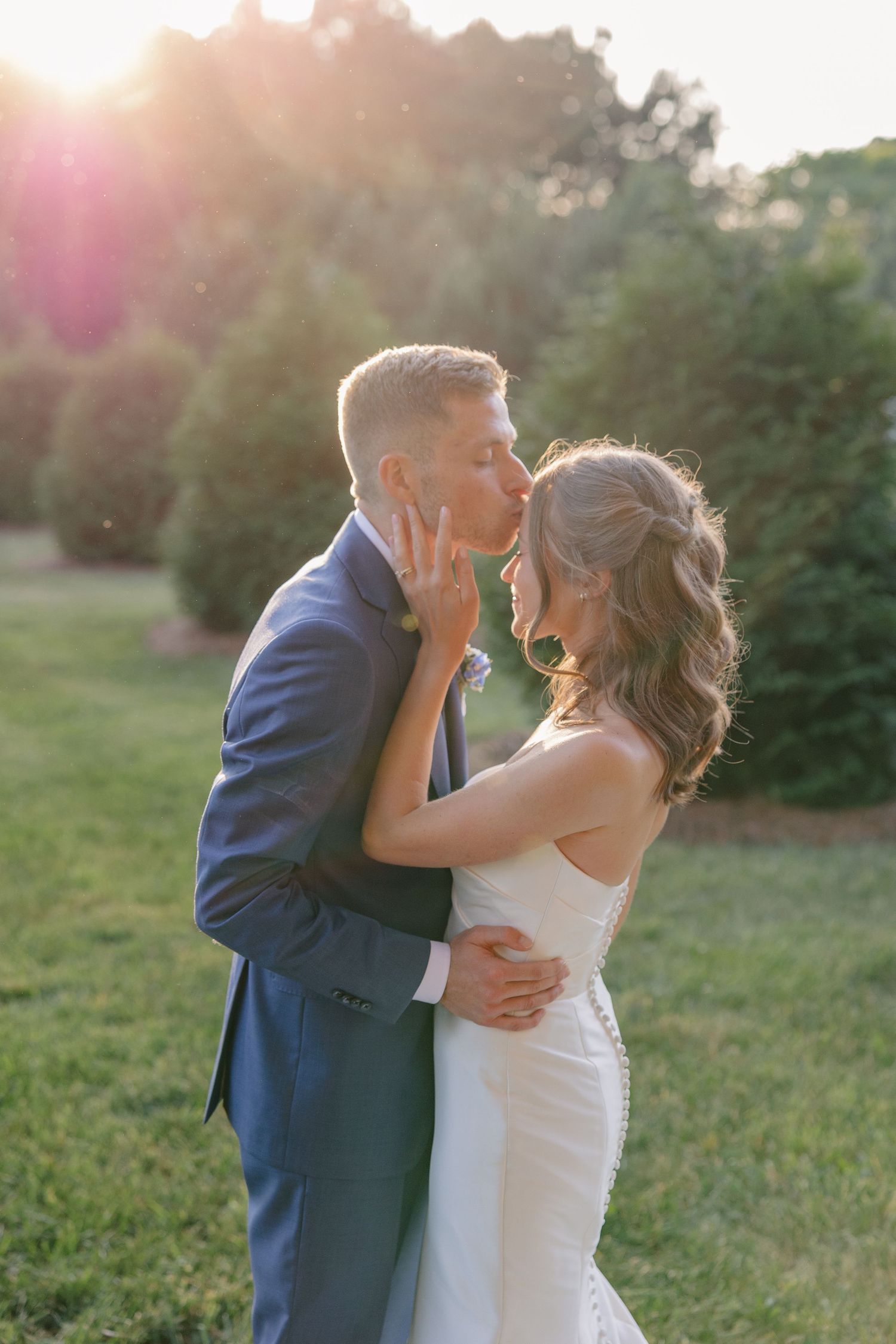 Bride and groom sharing a romantic kiss at golden hour sunset surrounded by lush green trees.