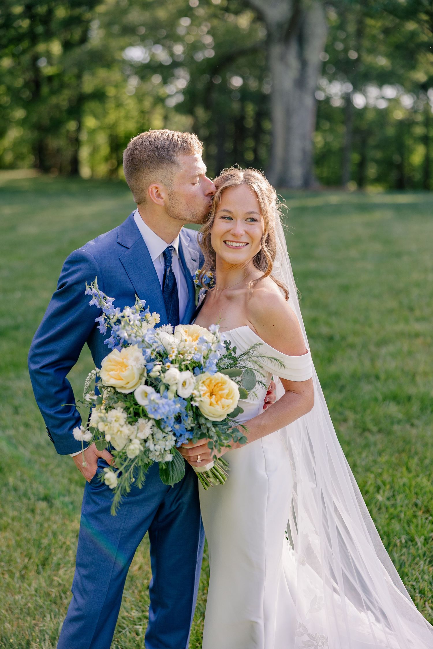 Groom in blue suit kisses bride on cheek as she smiles holding yellow and blue floral bouquet outdoors.