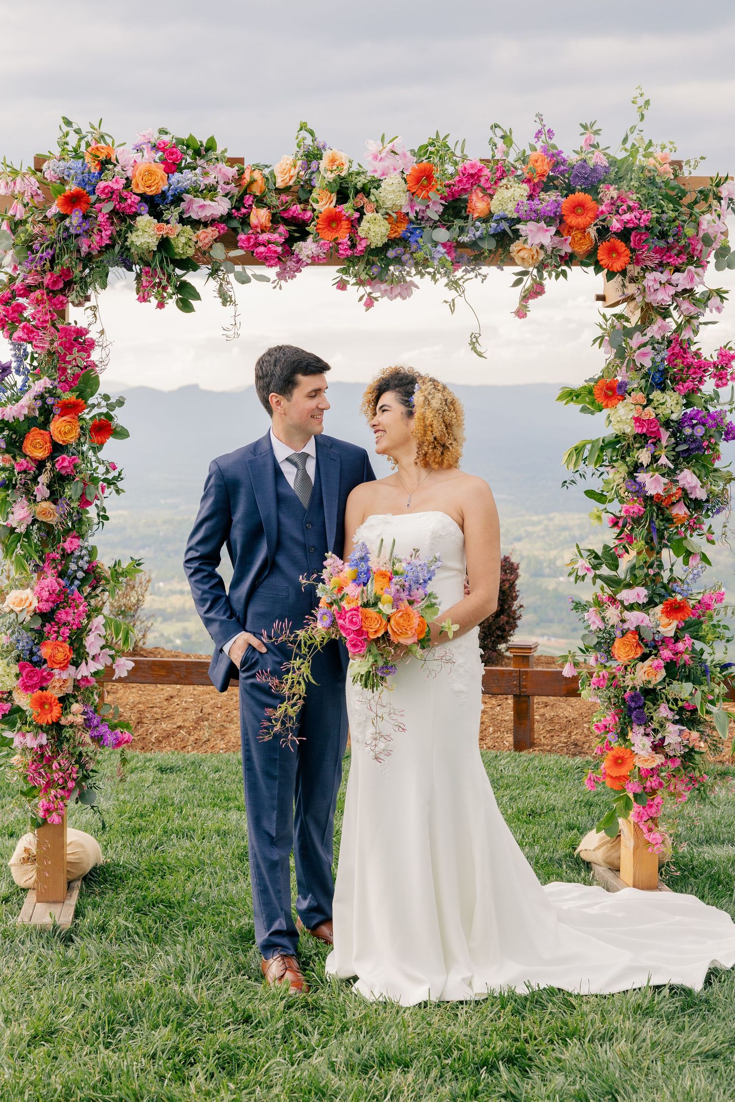 Happy couple sharing a romantic glance under a colorful flower arch with stunning mountain backdrop at their wedding.