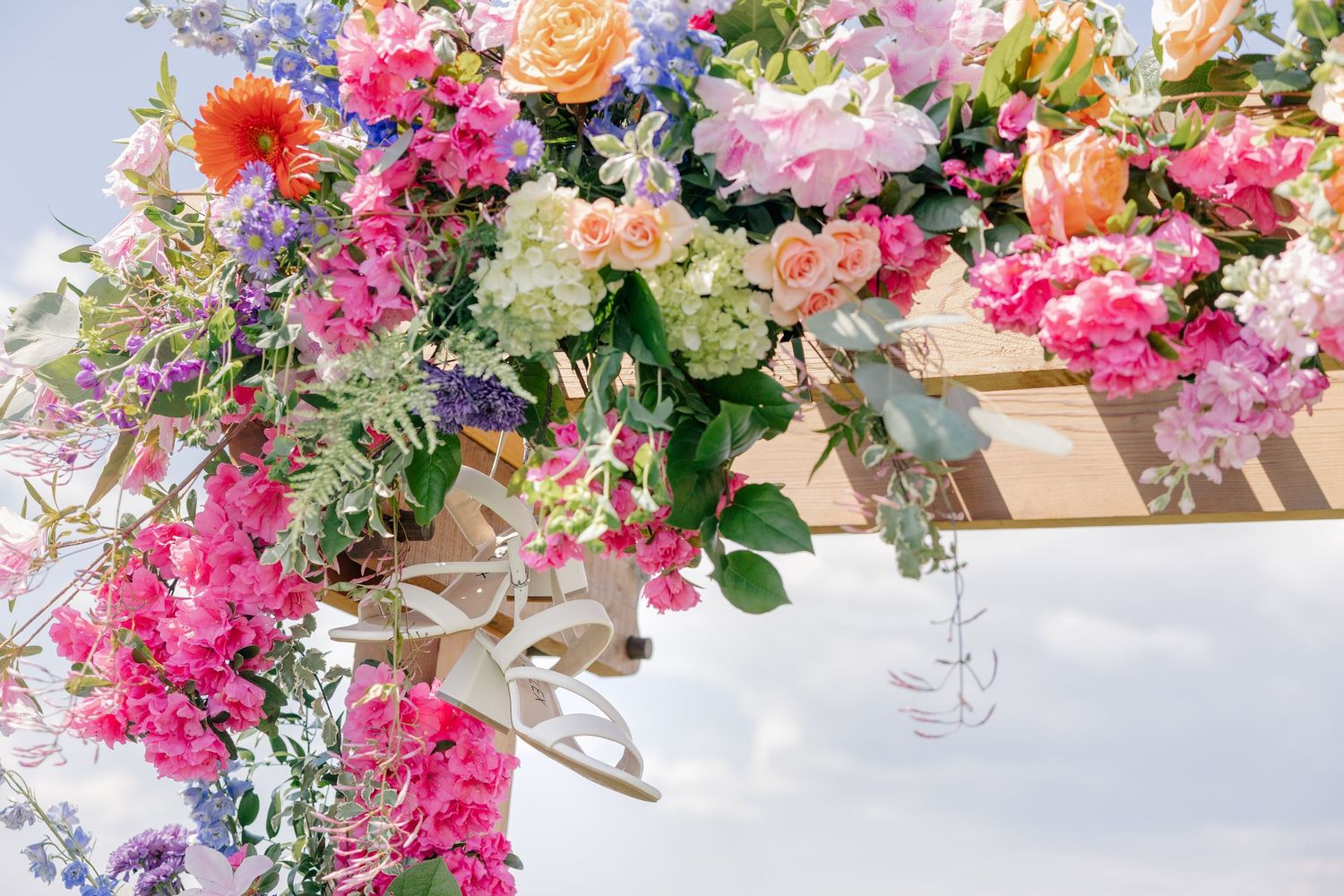 Colorful floral wedding arch adorned with pink, orange, and purple blooms against a bright sky.