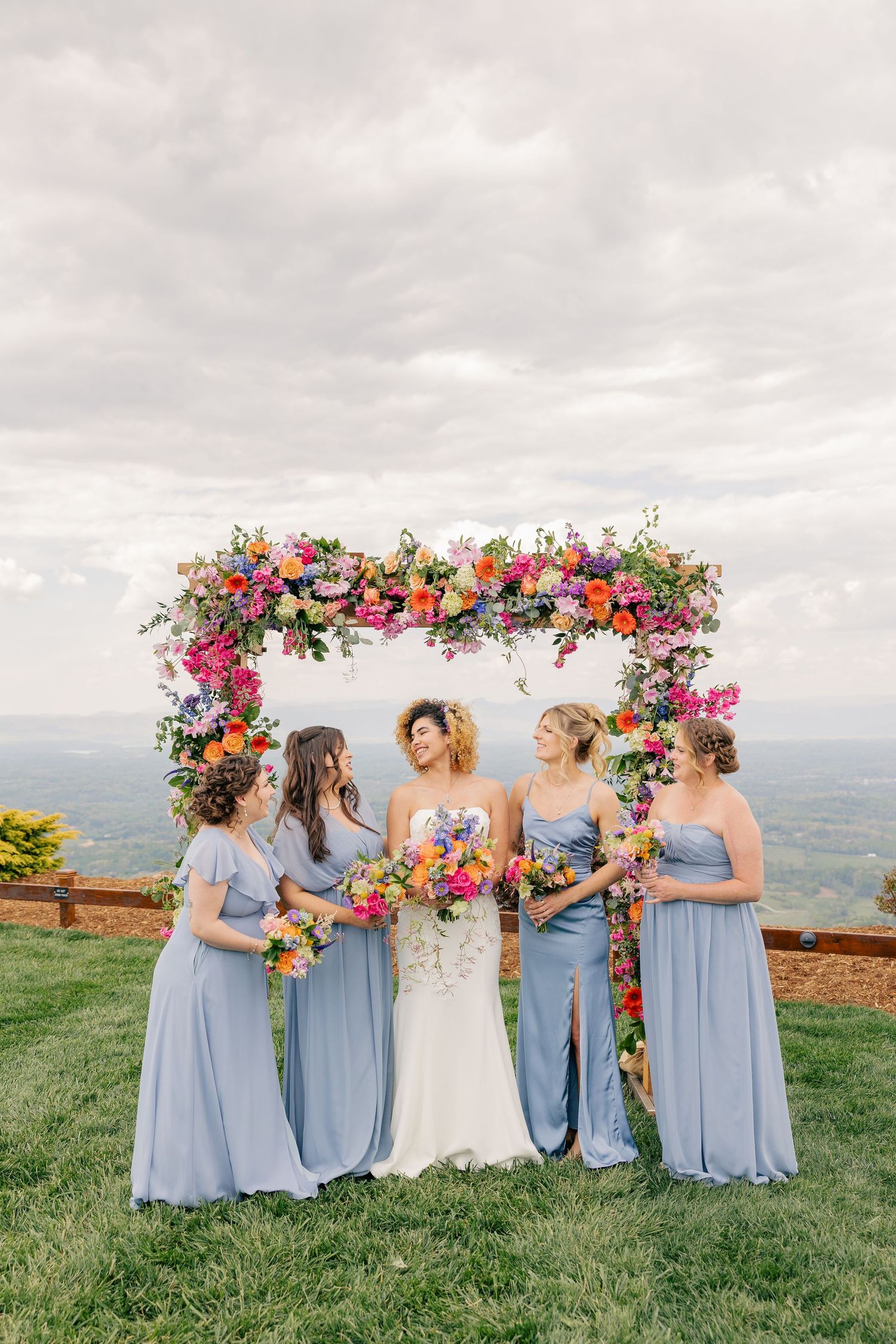 Bride in white gown stands with four bridesmaids in blue dresses under colorful floral arch with scenic view.