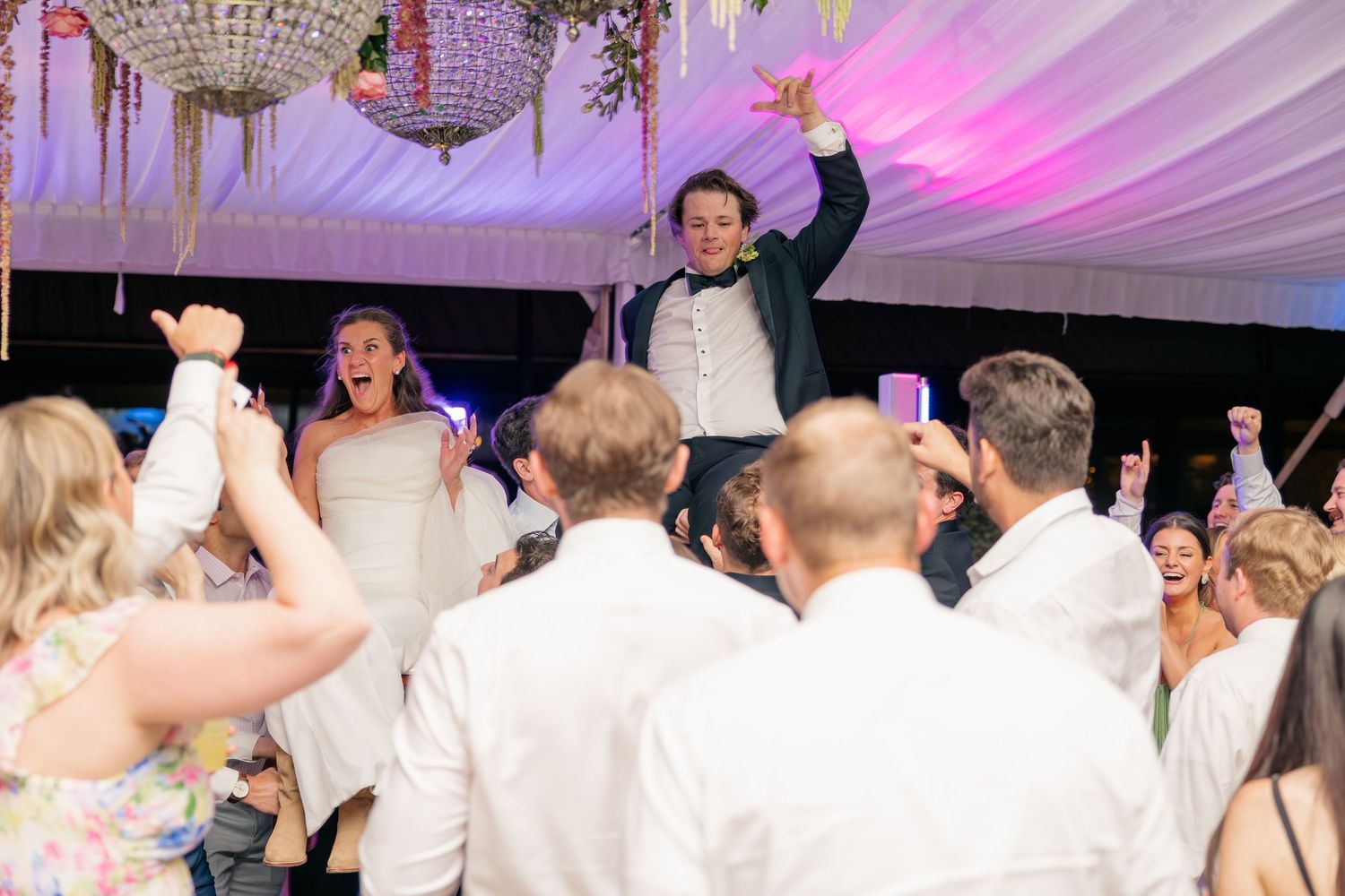 Joyful bride and groom lifted by celebrating guests on lively wedding reception dance floor.