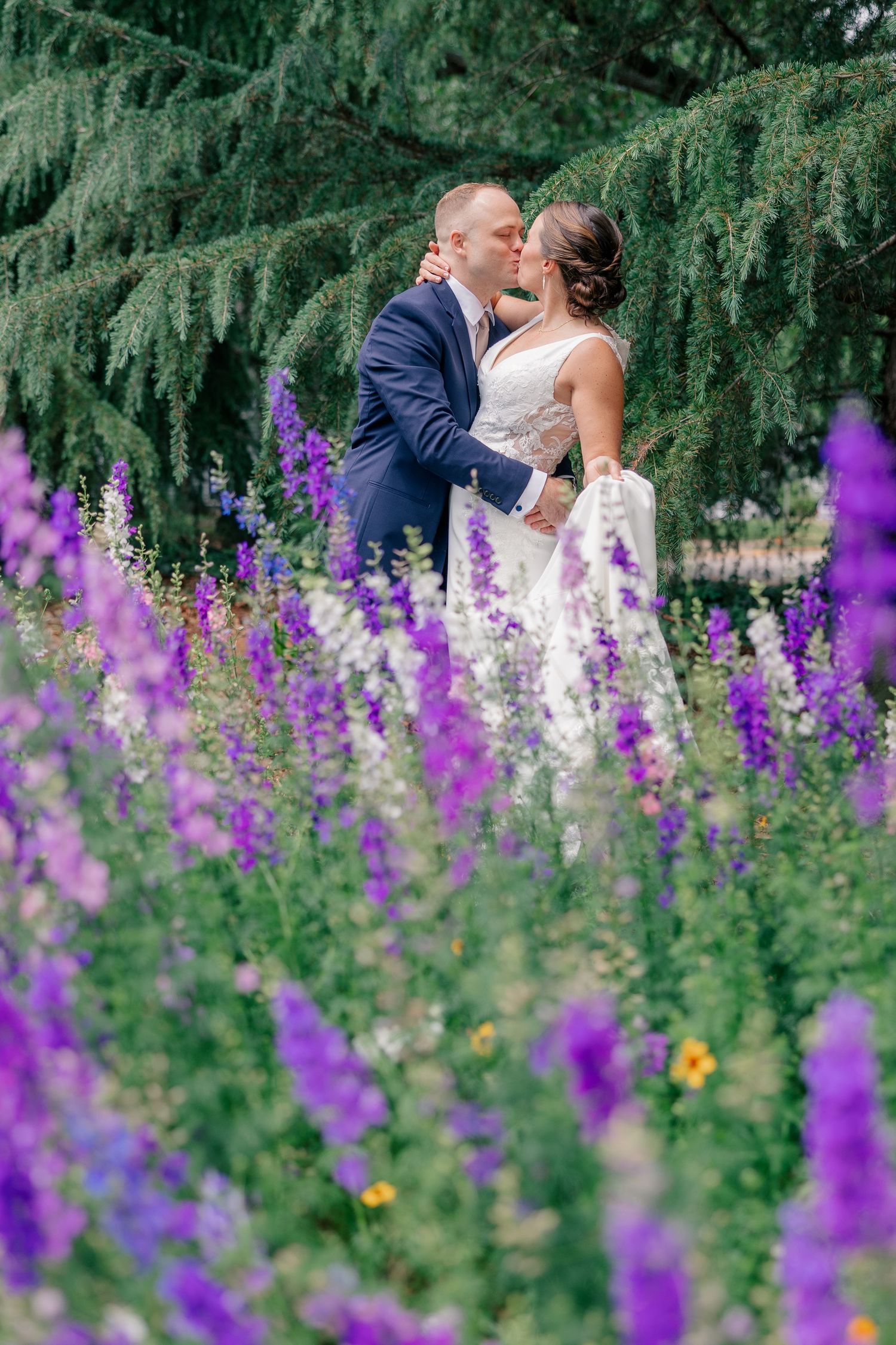 Bride and groom share a romantic kiss surrounded by vibrant purple wildflowers and evergreen trees.