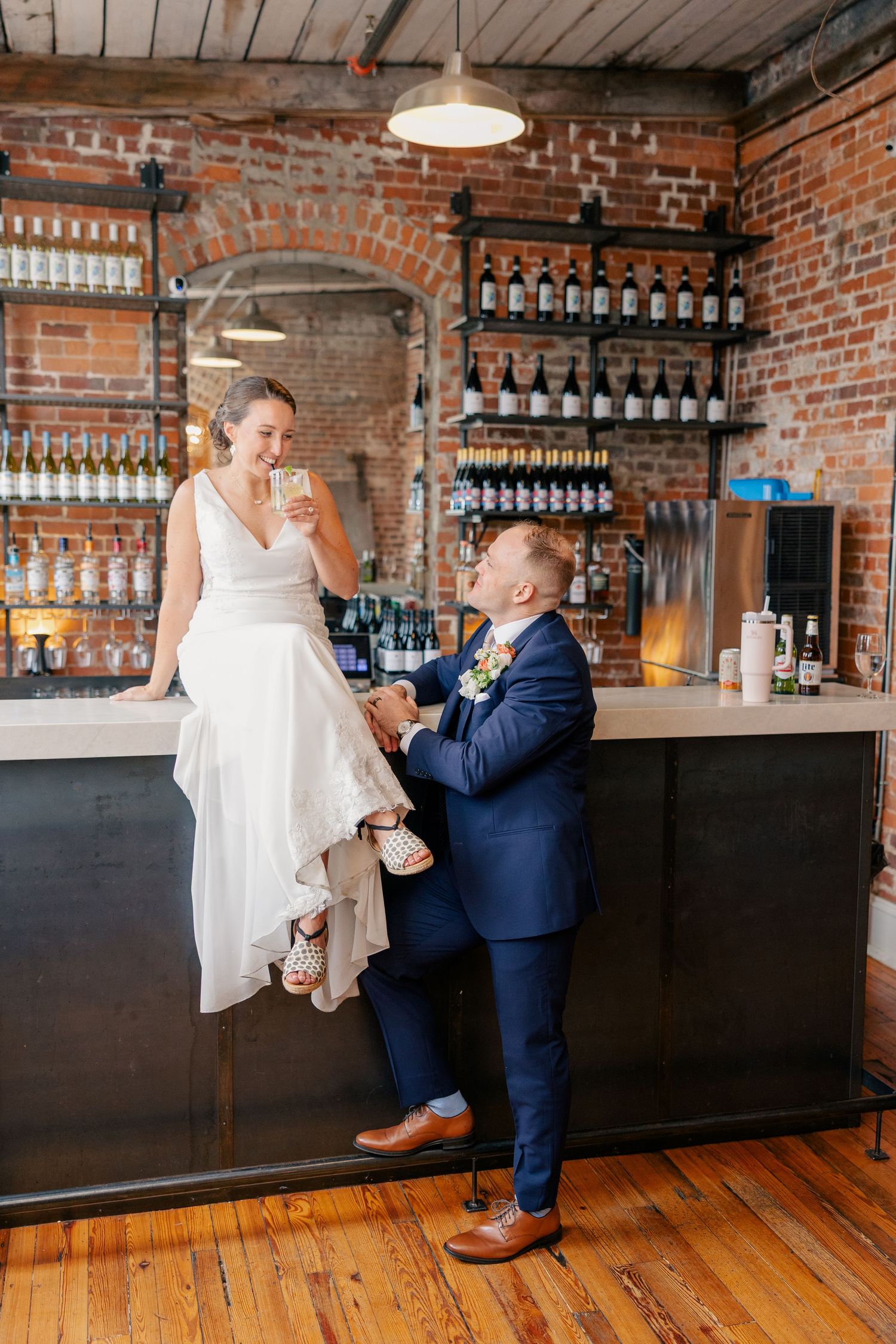Bride sitting on bar counter in white gown holding drink while groom in navy suit smiles at her in rustic venue.