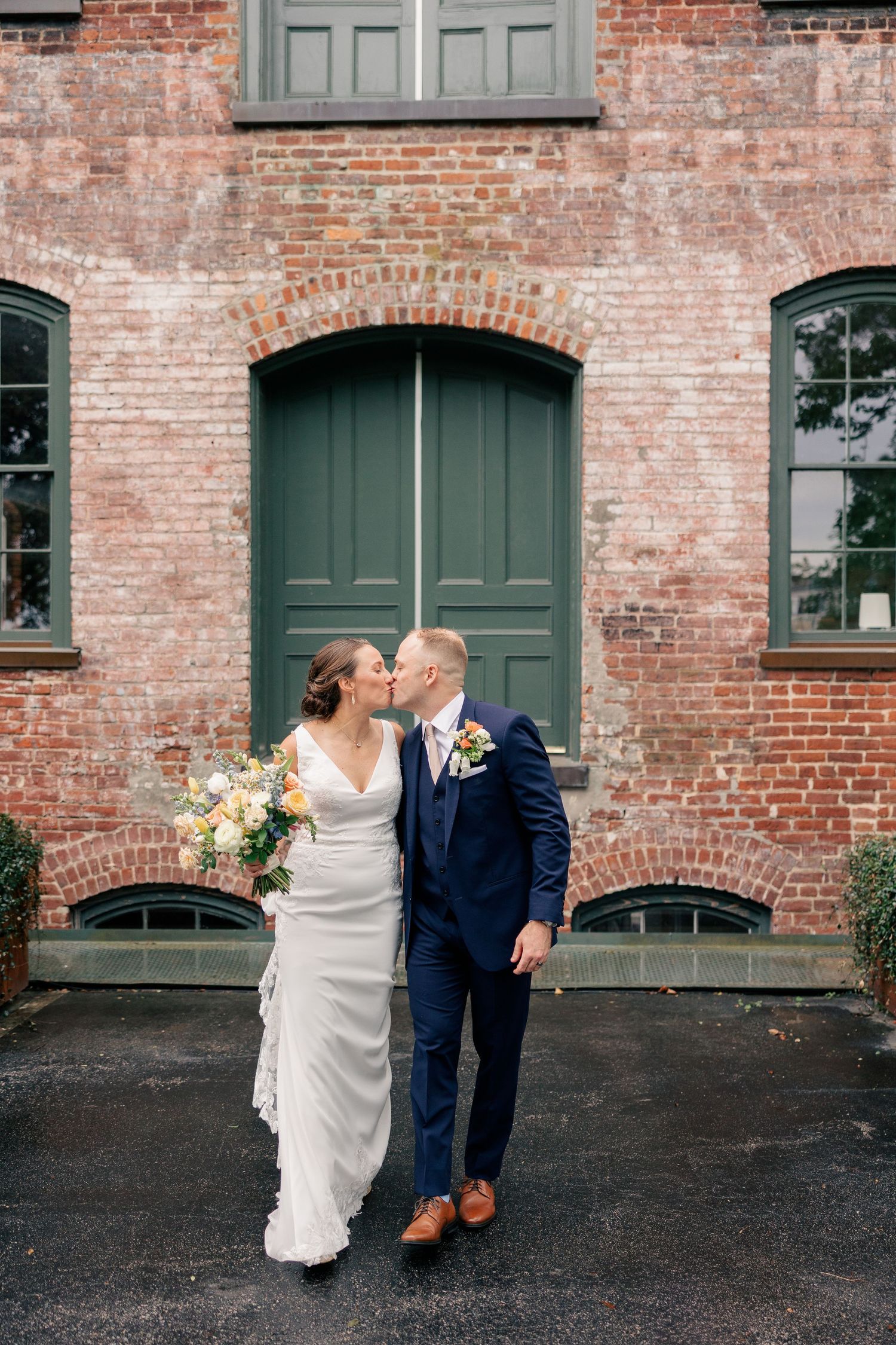 Bride and groom share a romantic kiss outside a rustic red brick building with green double doors.