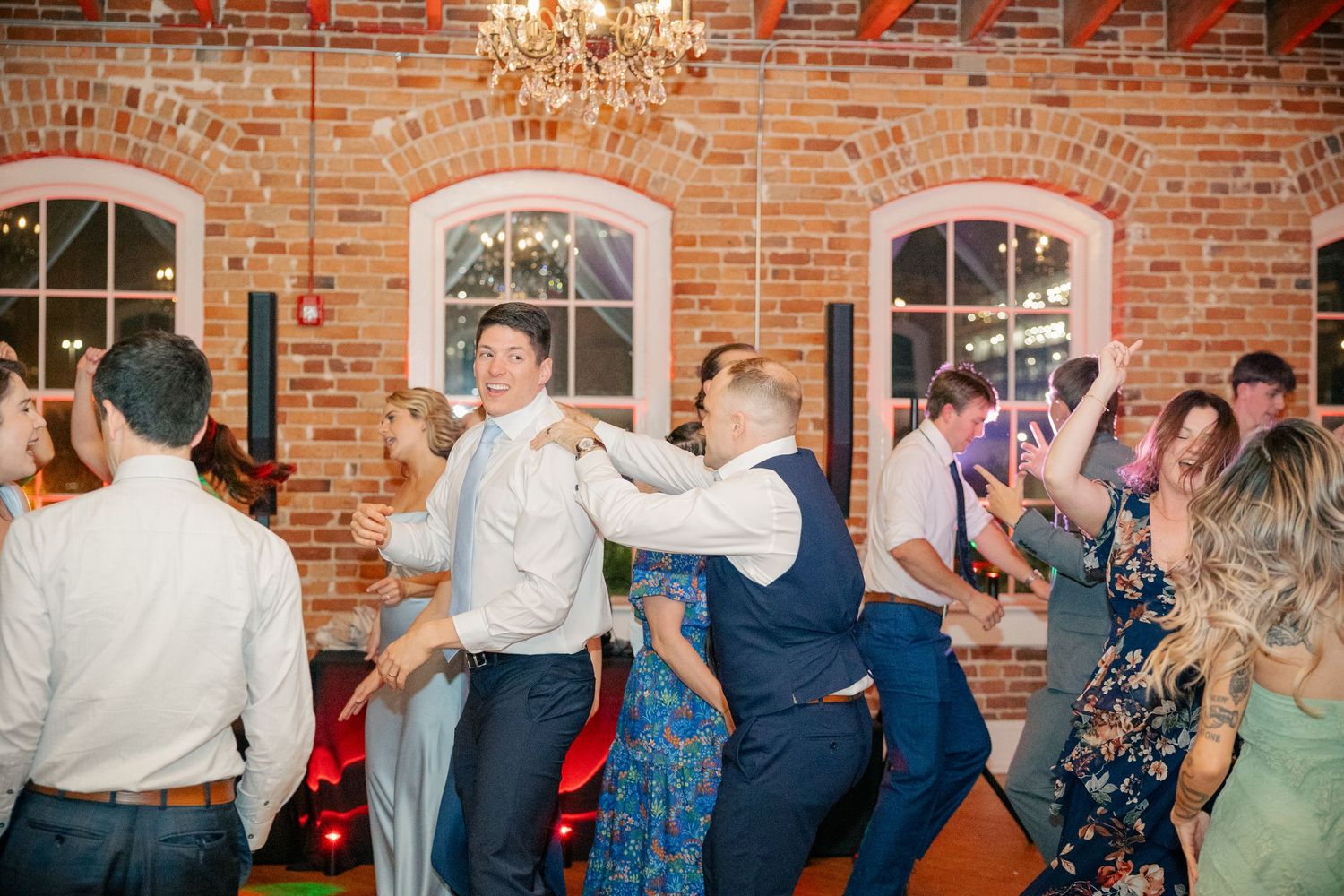 Wedding guests dance energetically together at lively reception in rustic brick venue with warm lighting.