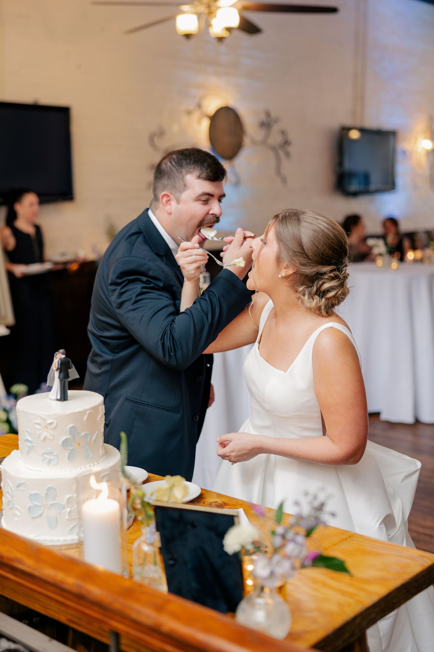Bride and groom laugh while feeding each other wedding cake at elegant indoor reception celebration.
