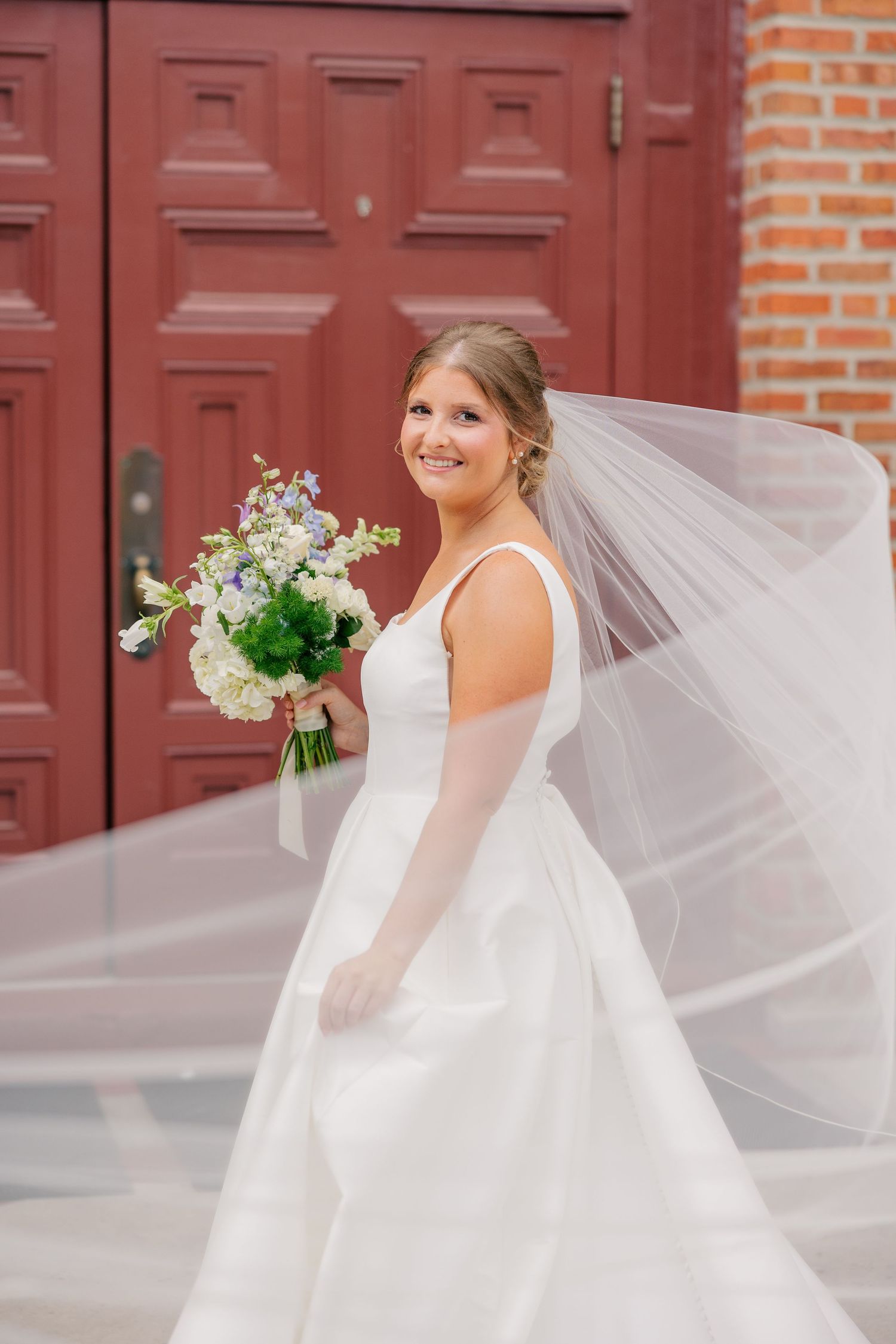 Smiling bride in sleeveless white gown holds blue and white bouquet with flowing veil at red doors.